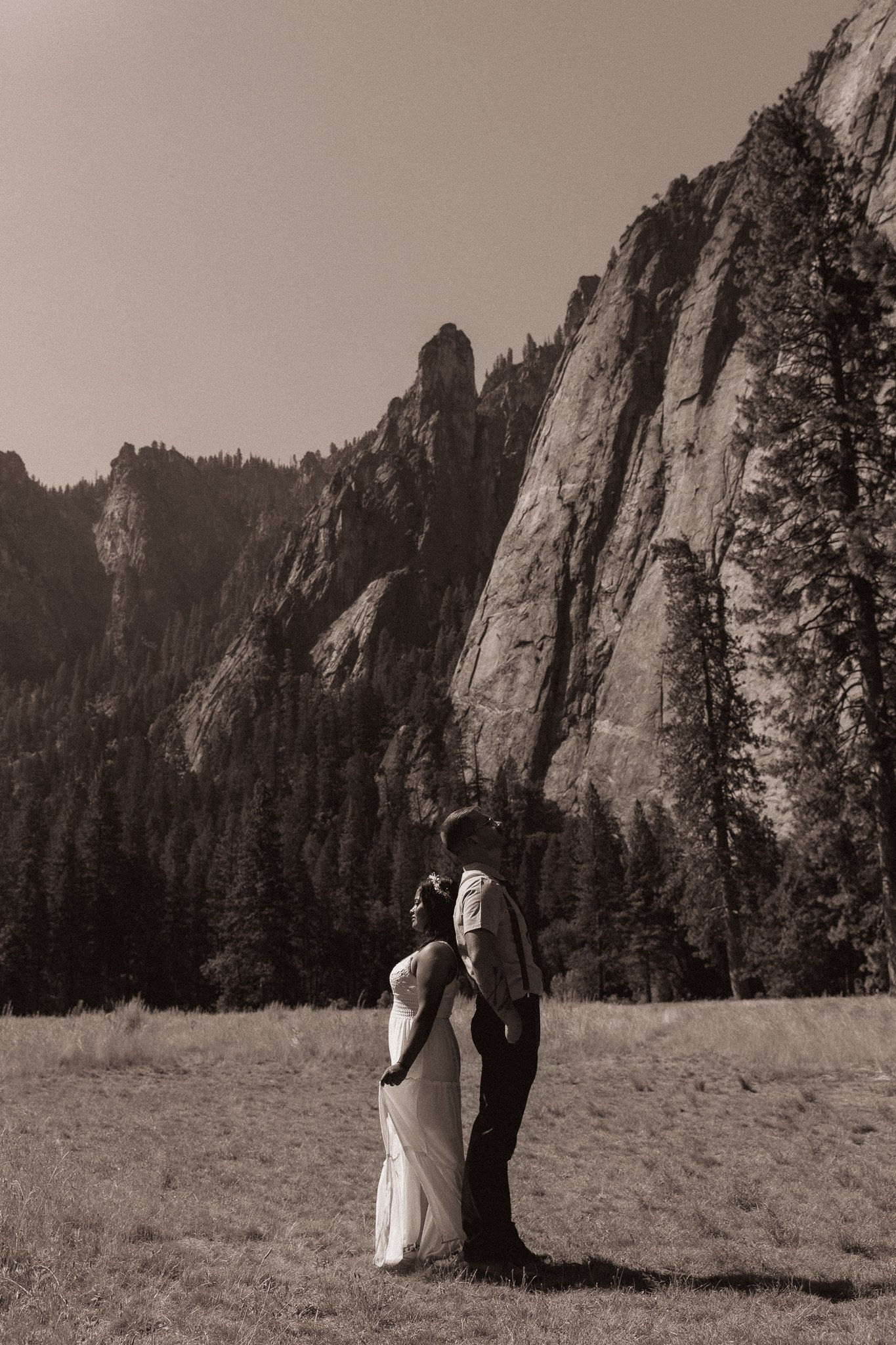 Couple standing back to back in a meadow with towering rocky cliffs and pine trees in the background, sepia tone.
