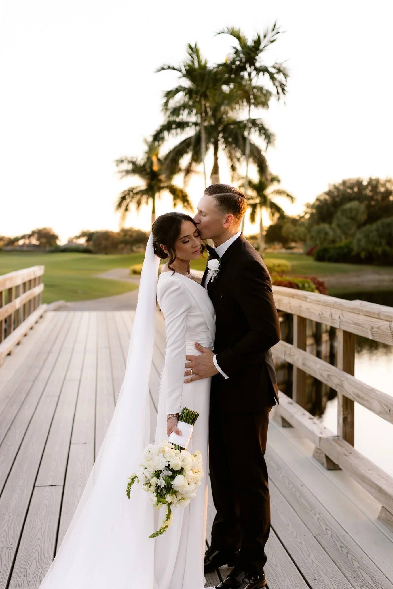 A bride and groom are embracing on a wooden bridge, with palm trees and a park in the background during sunset. The bride holds a bouquet of white roses, and the groom kisses her forehead.