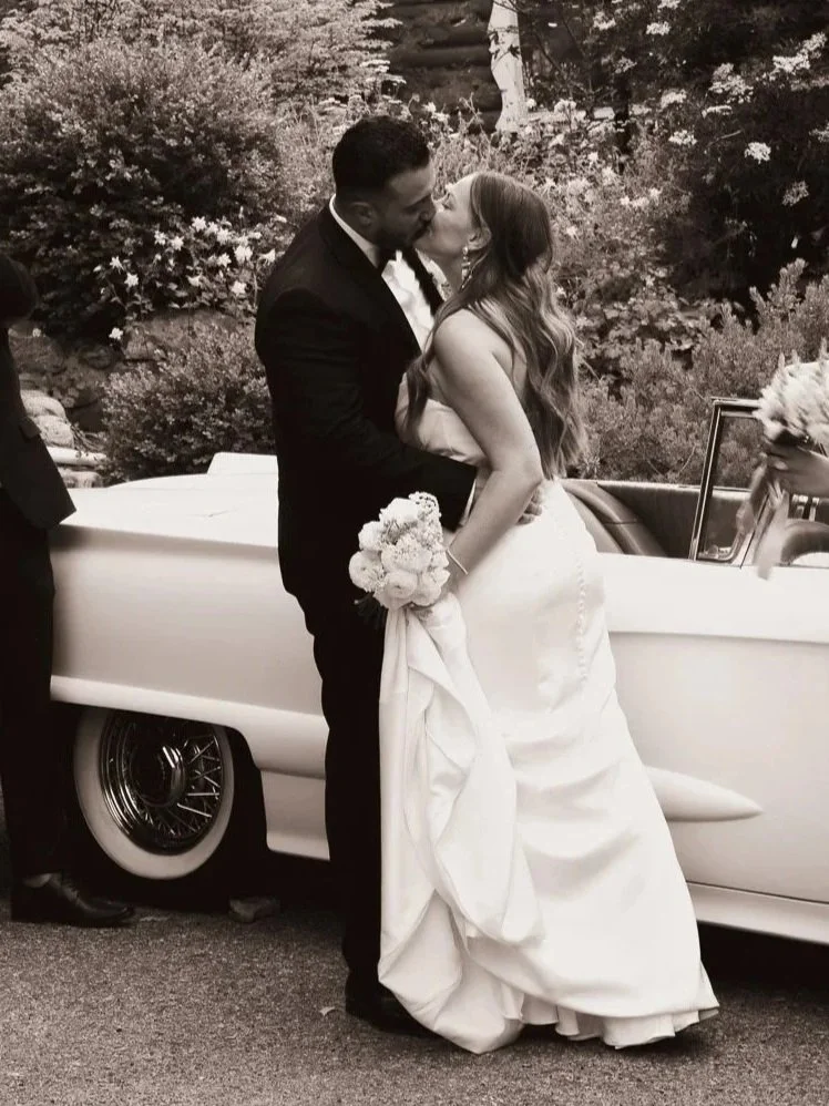 A newlywed couple sharing a kiss in front of a vintage car, with the bride holding a bouquet of flowers.