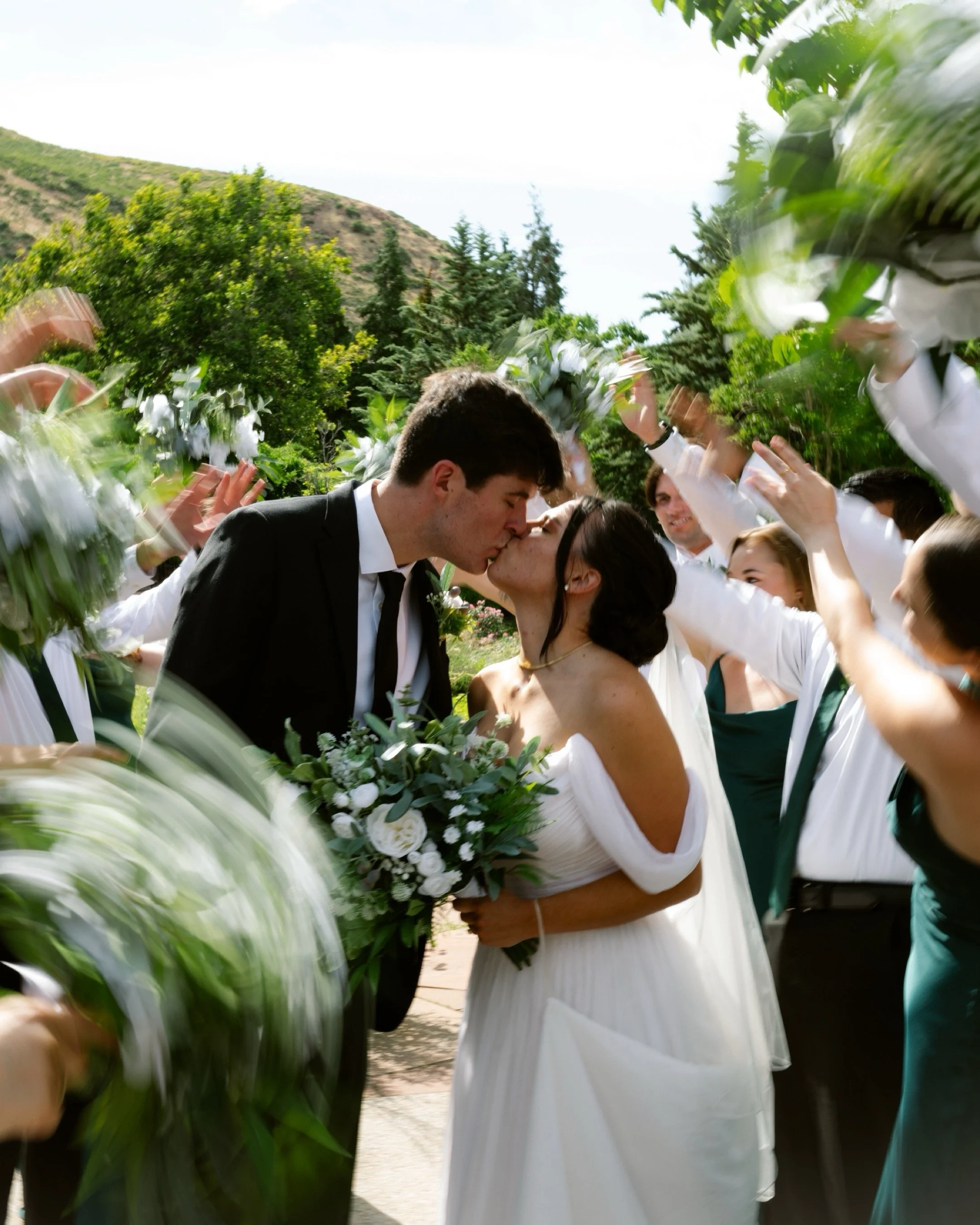 A couple dressed in wedding attire sharing a kiss during their outdoor wedding ceremony, surrounded by friends and family holding bouquets and celebrating.