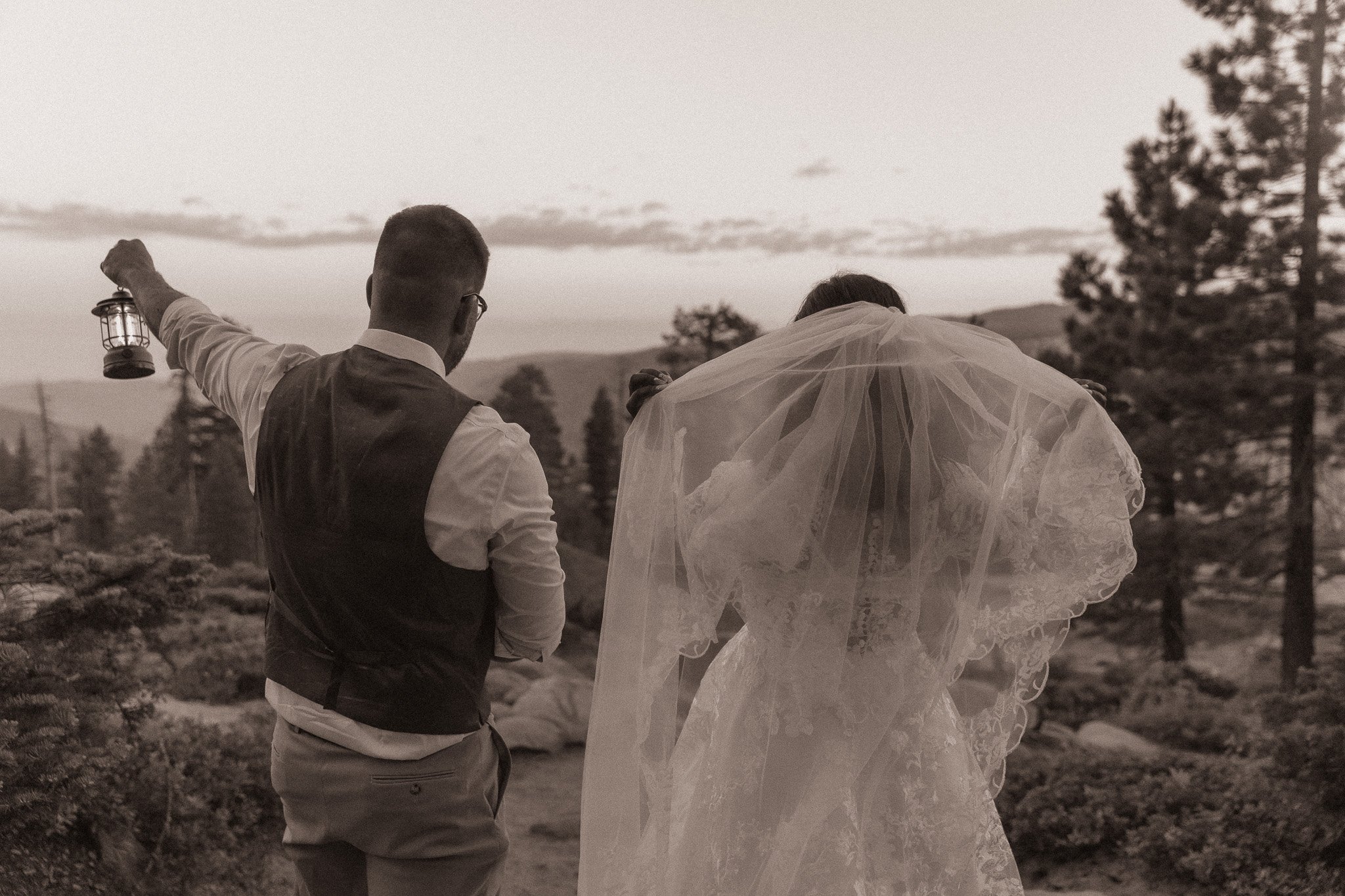 Bride and groom facing away, standing outdoors with groom holding a lantern, surrounded by trees and hills, in sepia tone.