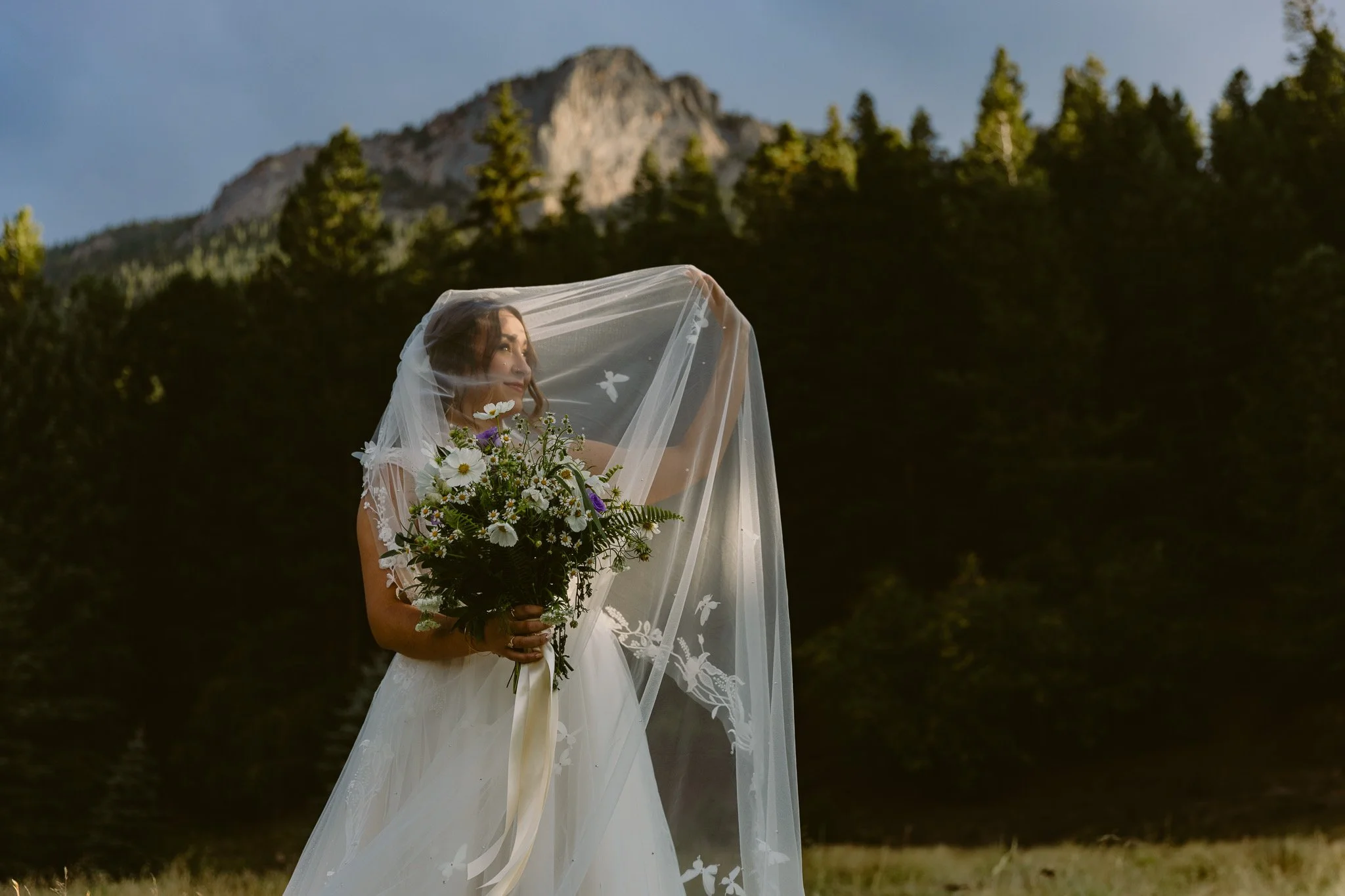A bride in a white wedding dress holding a bouquet of white and purple flowers, standing outdoors with a mountain and trees in the background, partially covered with a sheer veil.
