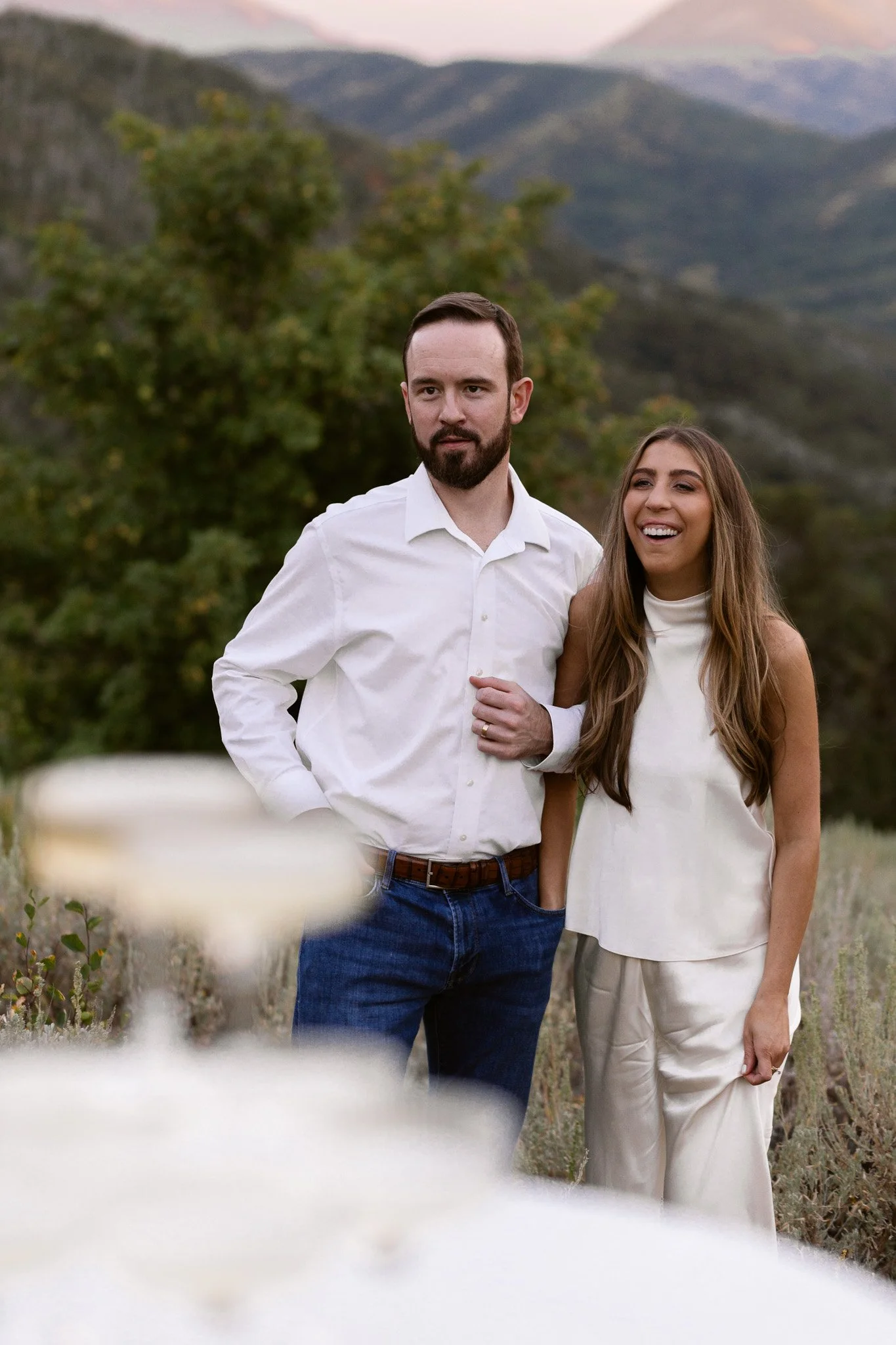 A man with dark hair and a beard wearing a white button-up shirt and jeans, and a woman with long light brown hair wearing a sleeveless white blouse and beige pants, standing outdoors in a natural setting with mountains and trees in the background, s