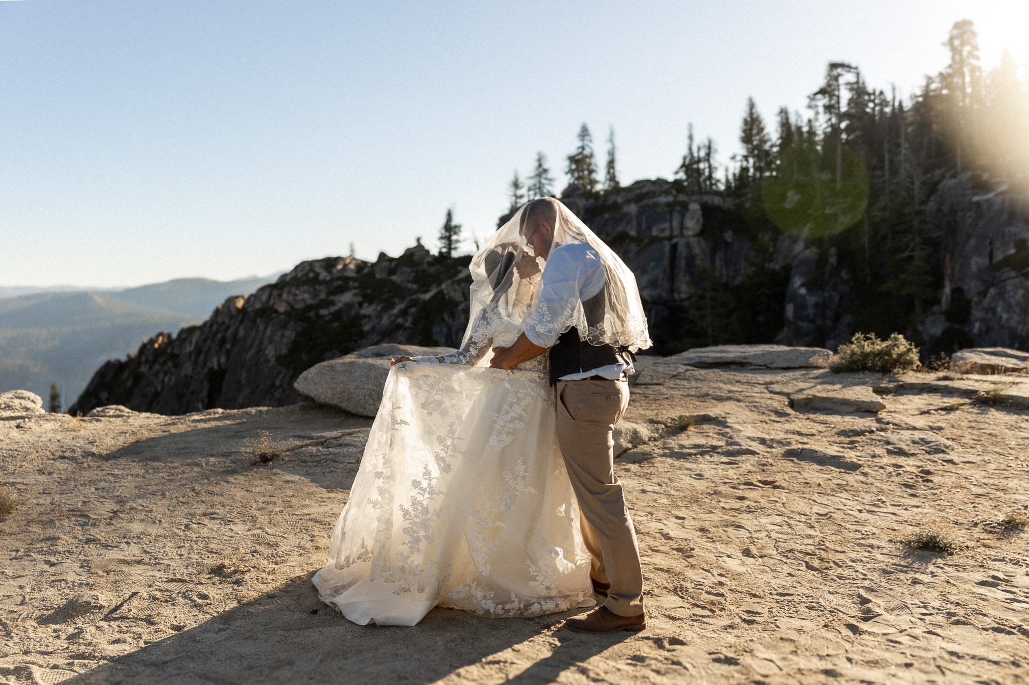 Bride and groom under veil on mountain top, embracing in natural setting with trees and rocks.