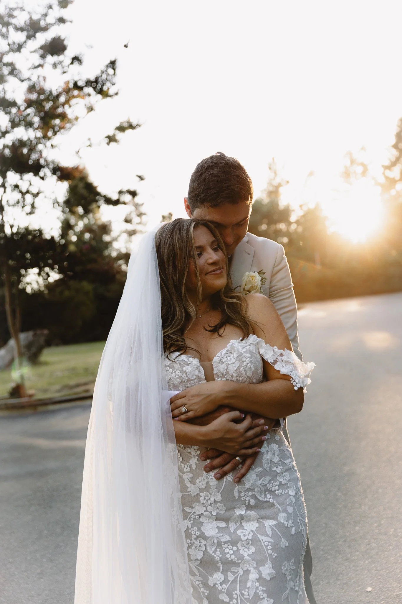 A bride in a white lace wedding dress and a groom in a white suit embrace outdoors during sunset.
