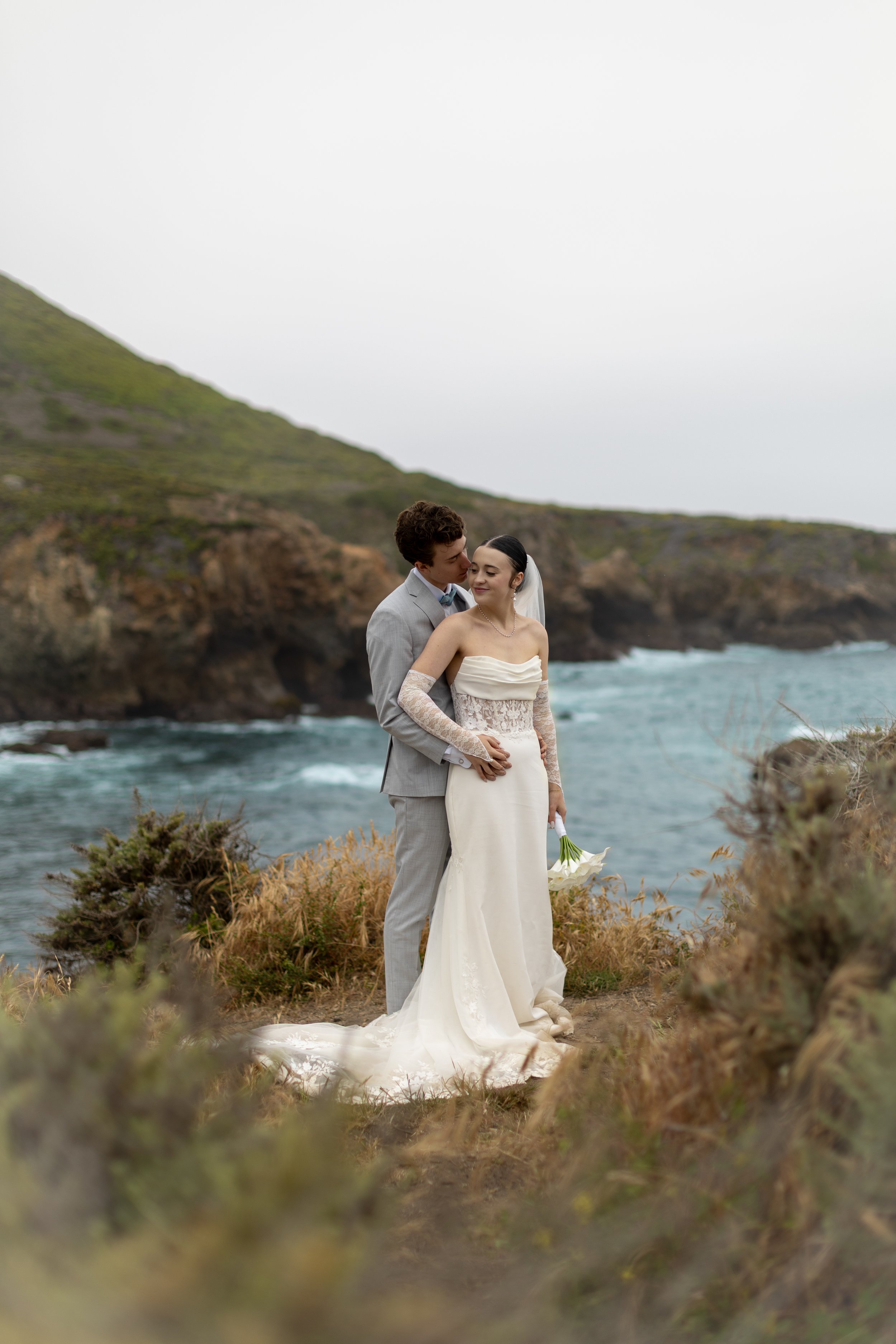 A bride and groom embrace on a rocky cliff near the ocean, with green hills and cloudy sky in the background.