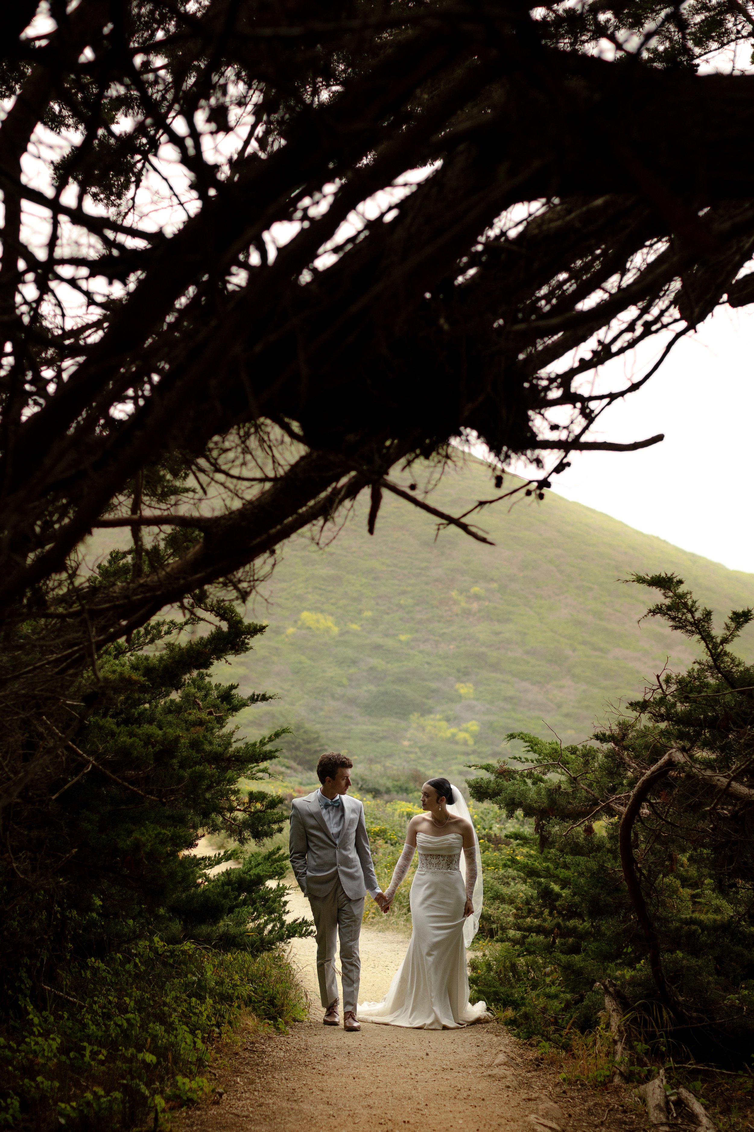 A bride and groom holding hands and walking along a trail in a forested area with a mountain in the background.