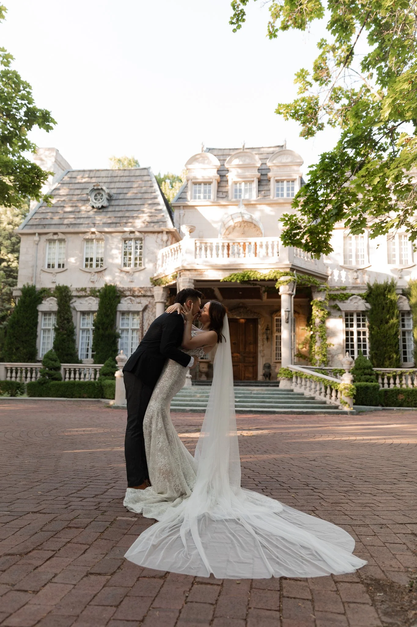A bride and groom share a kiss in front of a large, La Caille on a brick driveway, surrounded by trees and greenery.