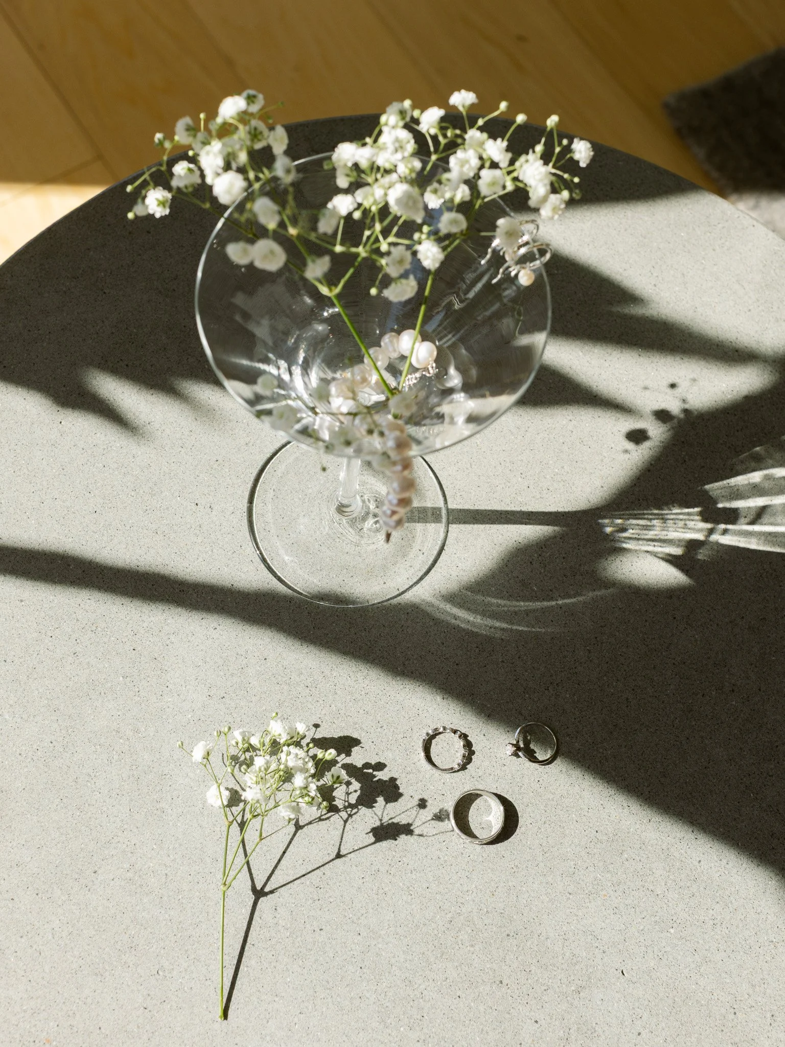 A top-down view of a gray table with a glass martini glass containing white baby's breath flowers, casting shadows. A single baby's breath flower and three rings are also on the table at Wind & Sea estate, Big Sur. 