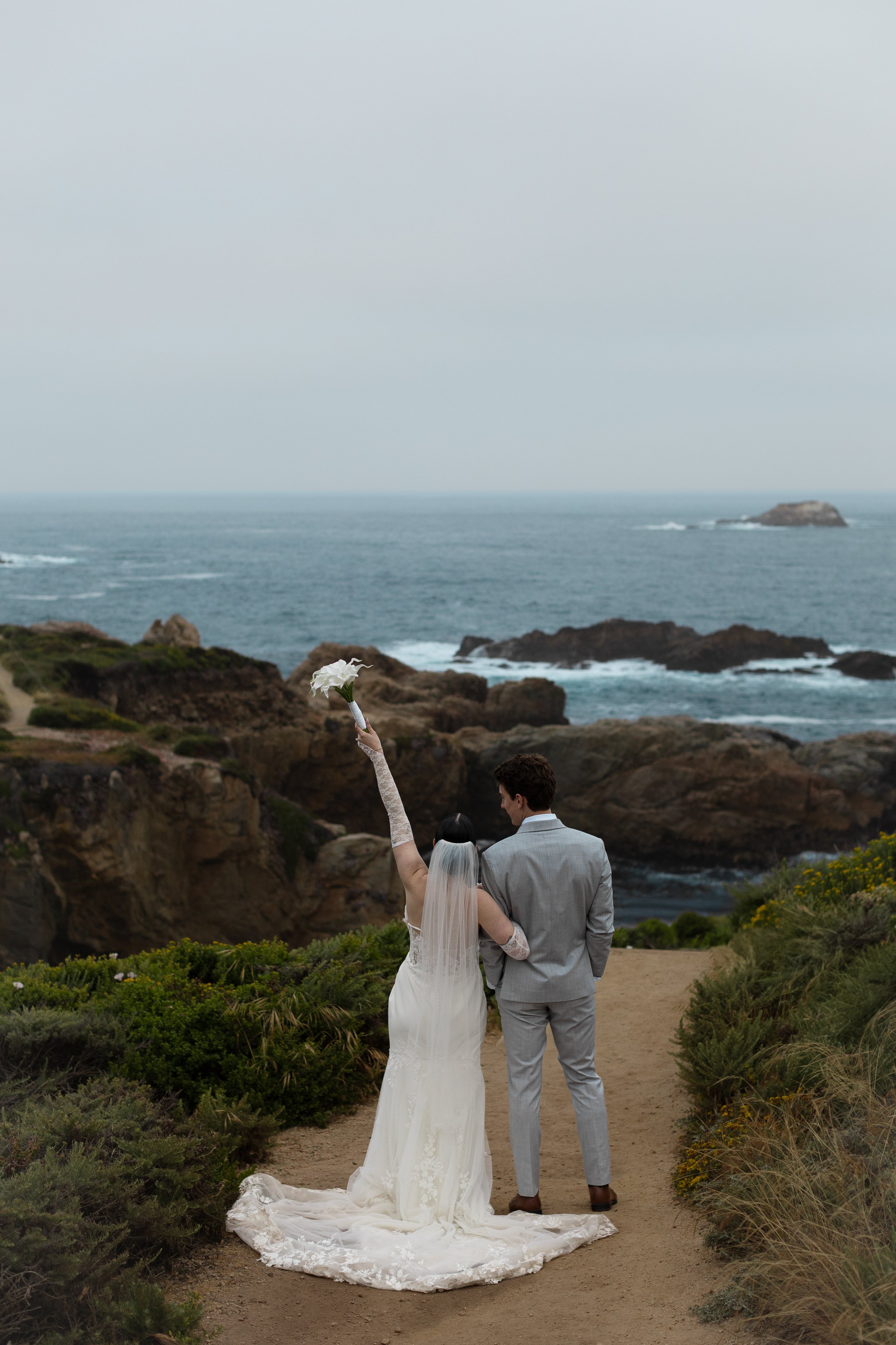 A bride and groom standing on a beach with the ocean in the background, the bride holding a bouquet above her head.