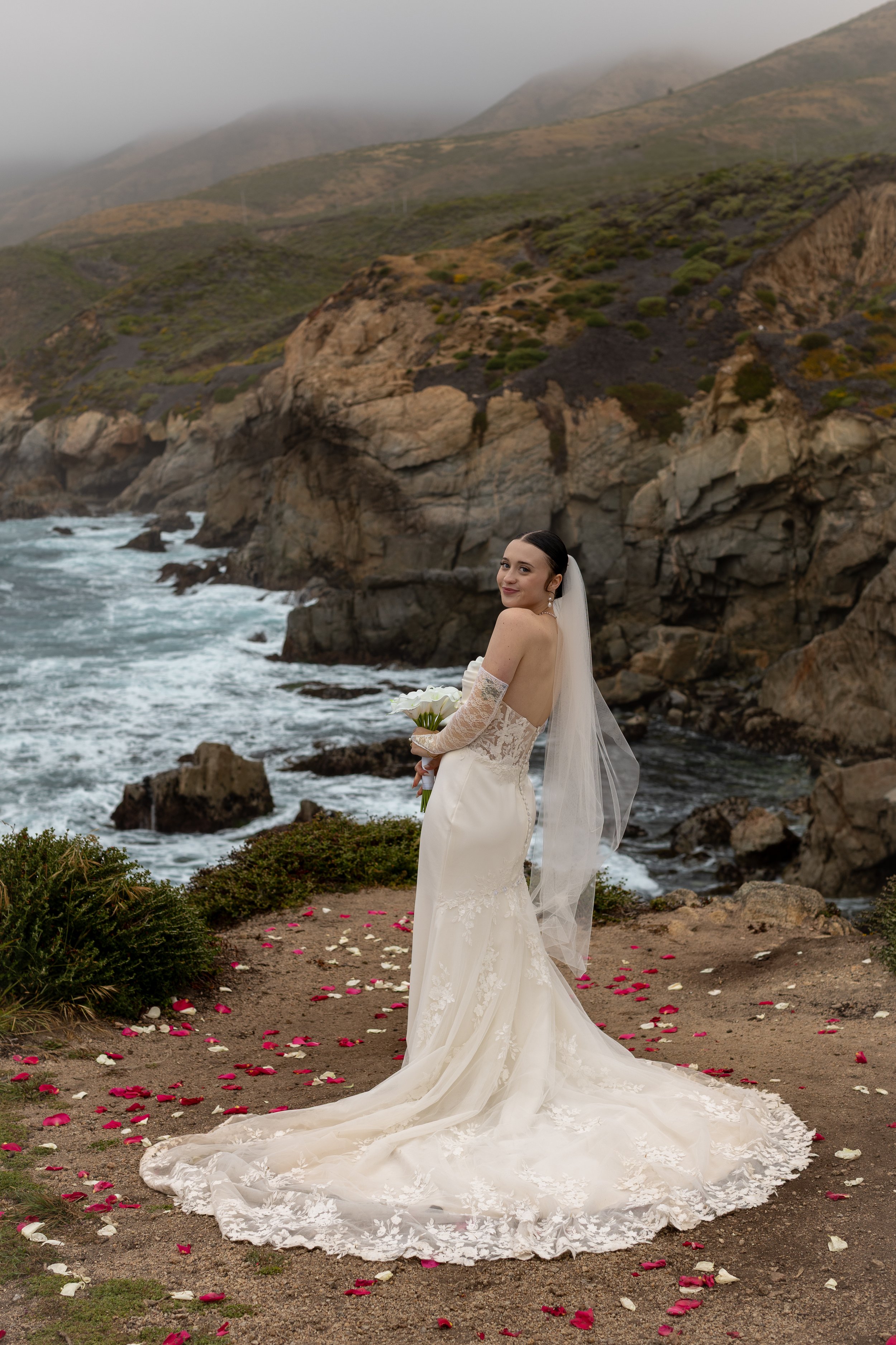 Bride in white wedding gown with lace sleeves holding a bouquet of white flowers, standing on a rocky coastal landscape with ocean waves and foggy mountains in the background, surrounded by scattered pink and white flower petals.