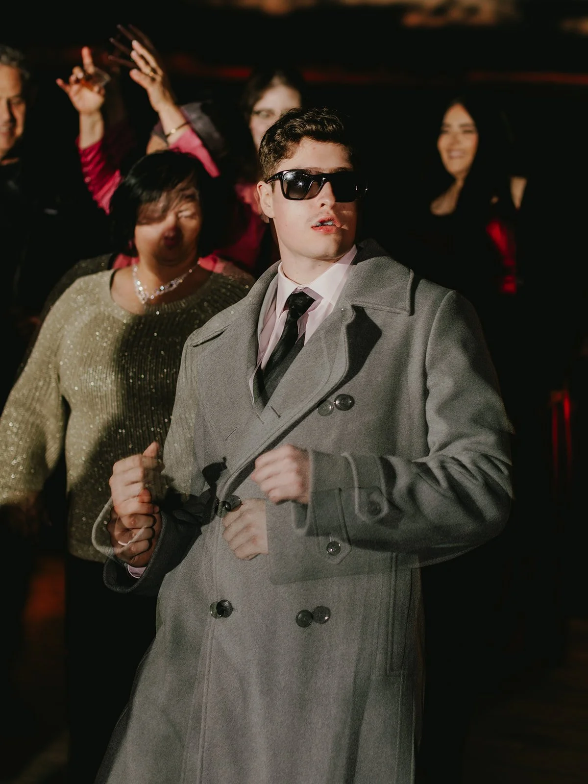 Young man wearing sunglasses, a gray trench coat, and a black tie, in a crowd at a party.