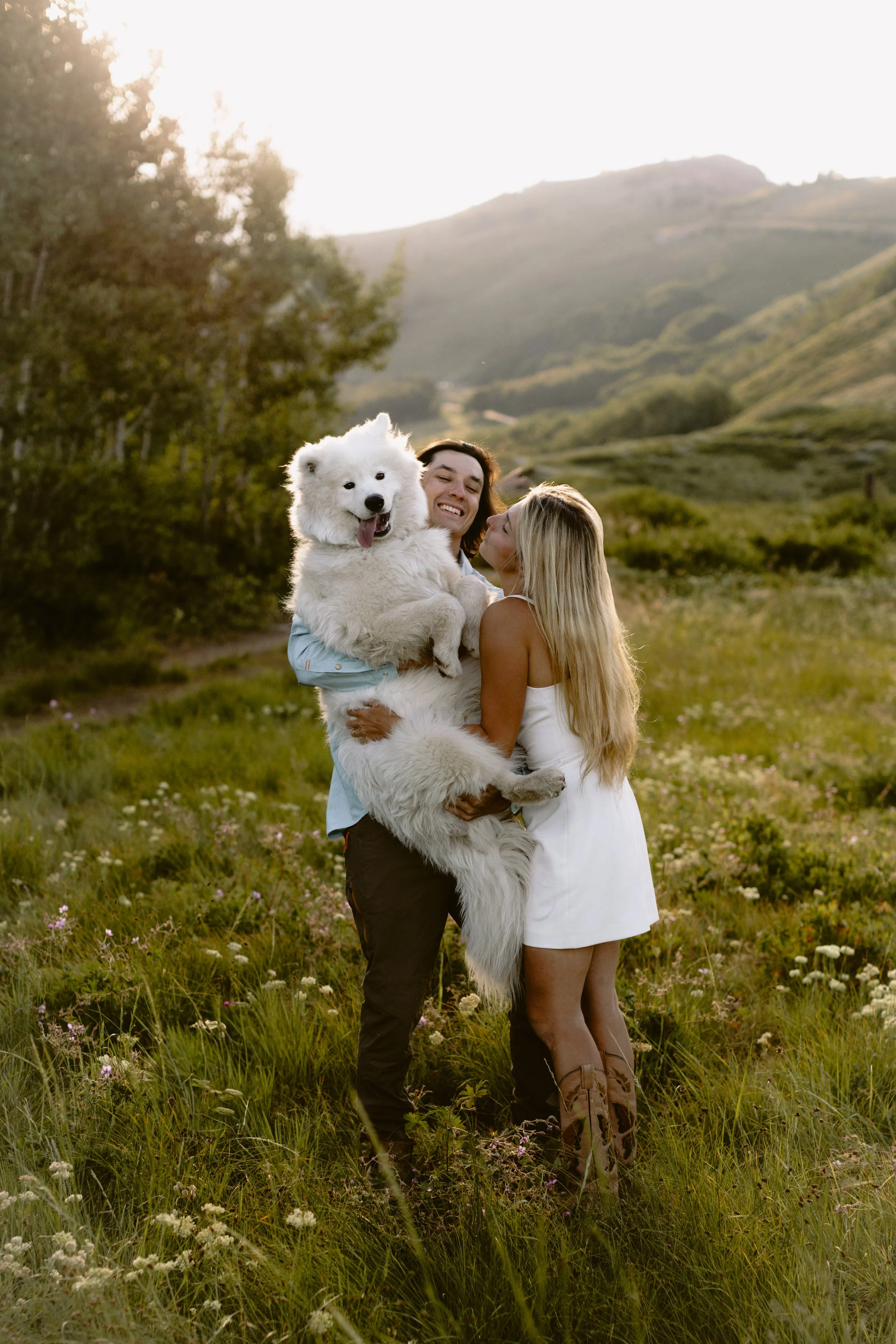 A happy couple embracing in a green field with a large white fluffy dog. The man is holding the dog, and the woman is kissing it, all three are smiling. The background features mountains and trees with sunlight shining from the left.