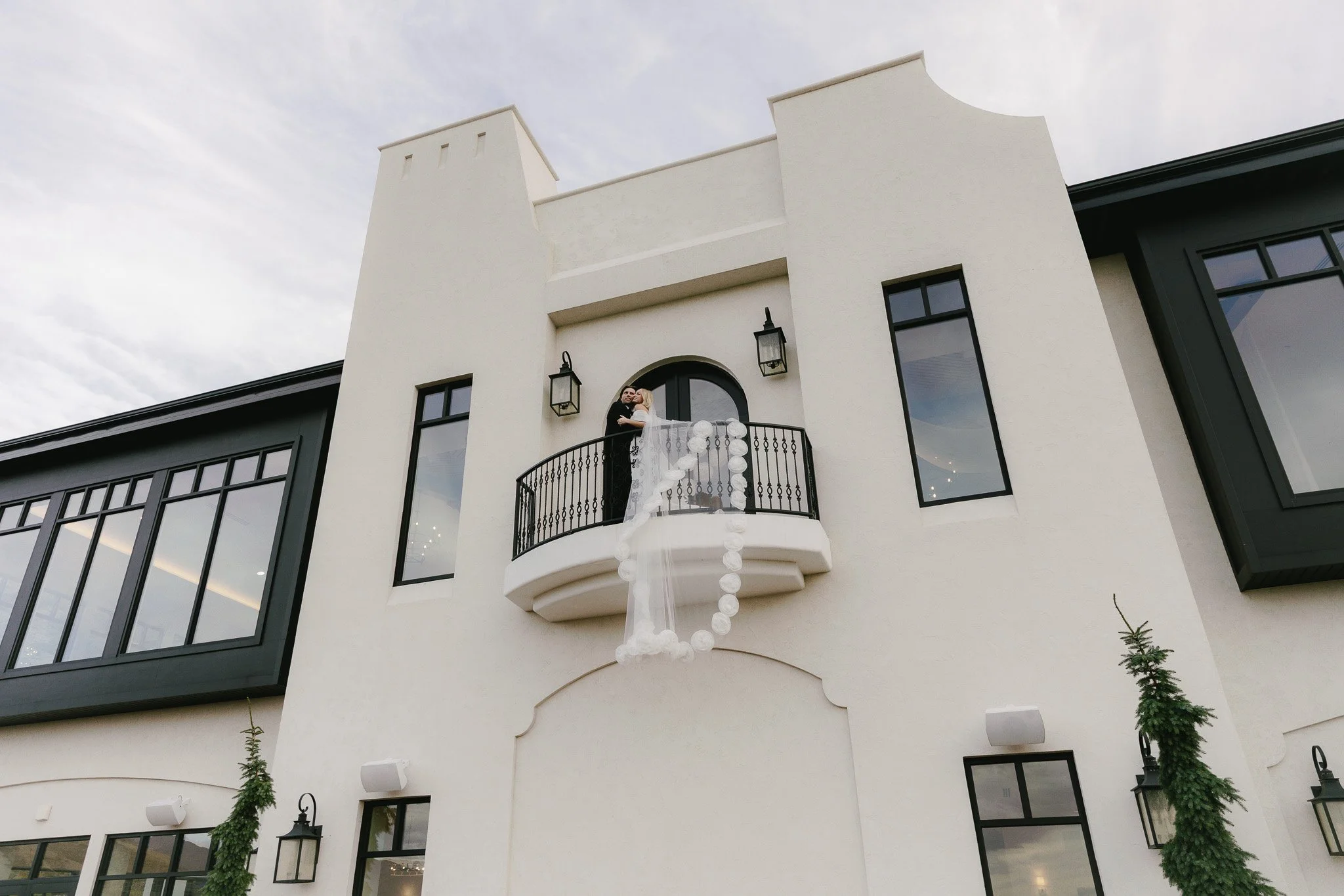A bride and groom dancing on a small balcony of a white building with black trim, outdoor lanterns, and tall windows.