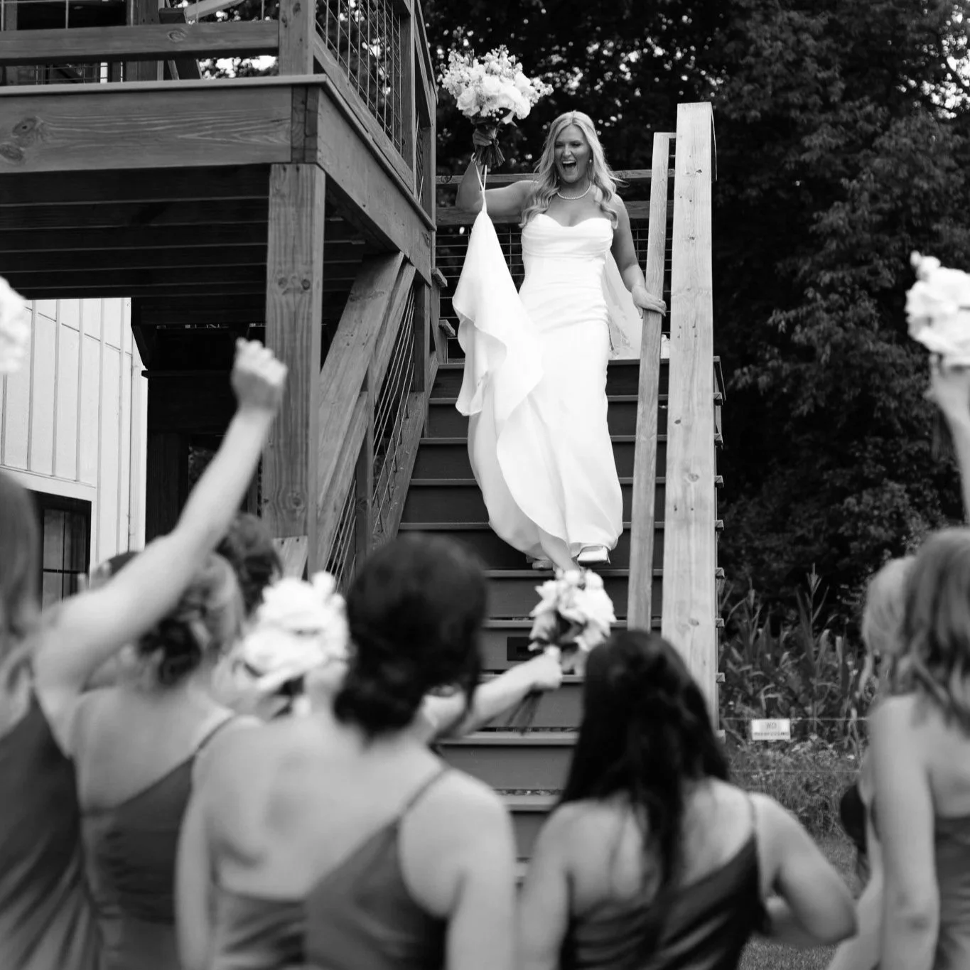 A bride in a white wedding dress walking down stairs holding a bouquet, smiling, with guests in the foreground cheering.