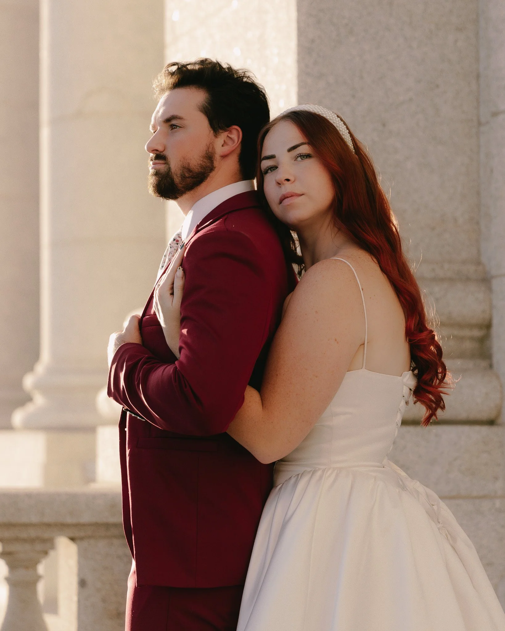 A man in a burgundy suit and a woman in a white dress are standing back-to-back outside near stone columns, with soft natural light illuminating their profiles.