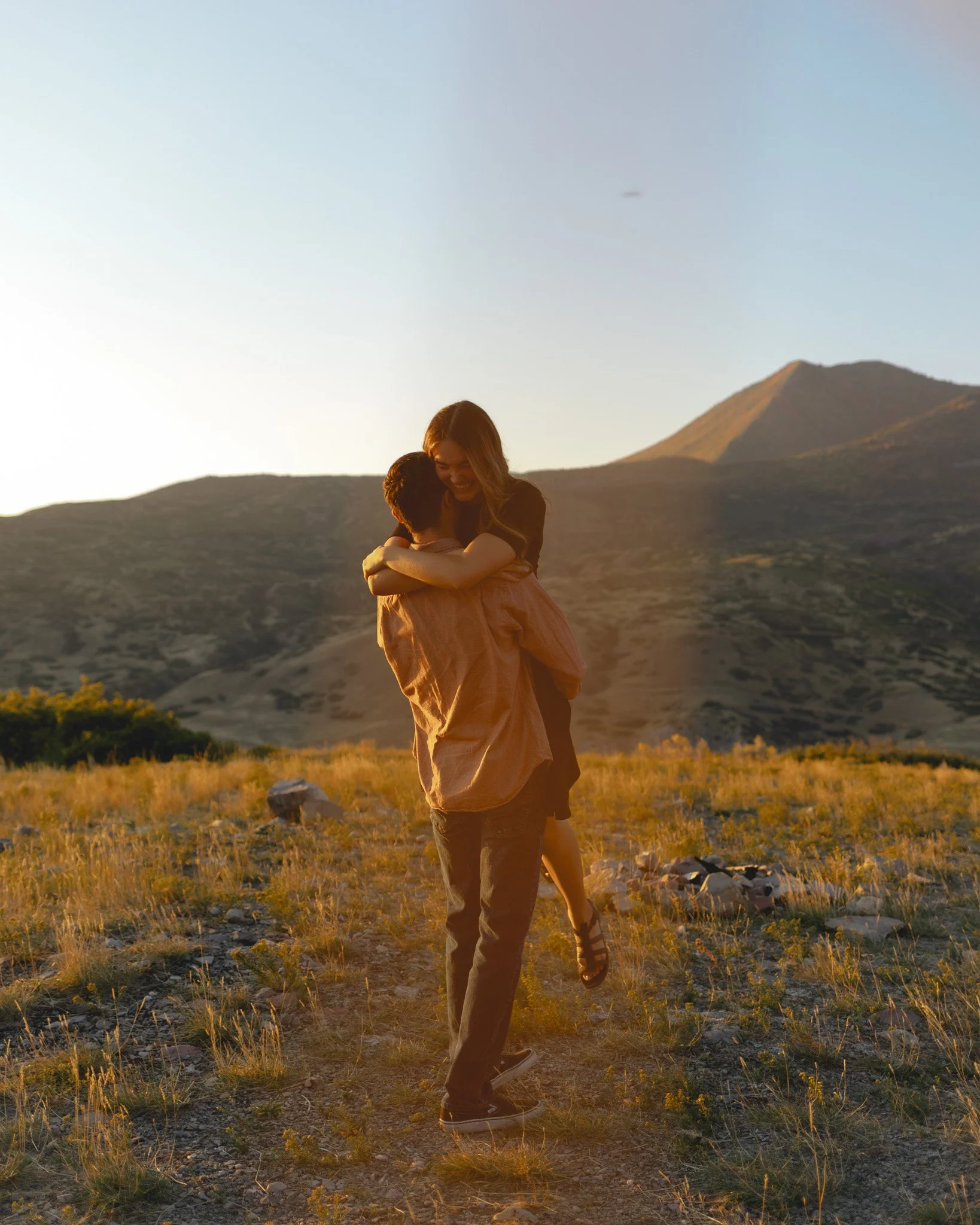 A woman is lifting and hugging a boy in a grassy outdoor setting during sunset, with mountains in the background.