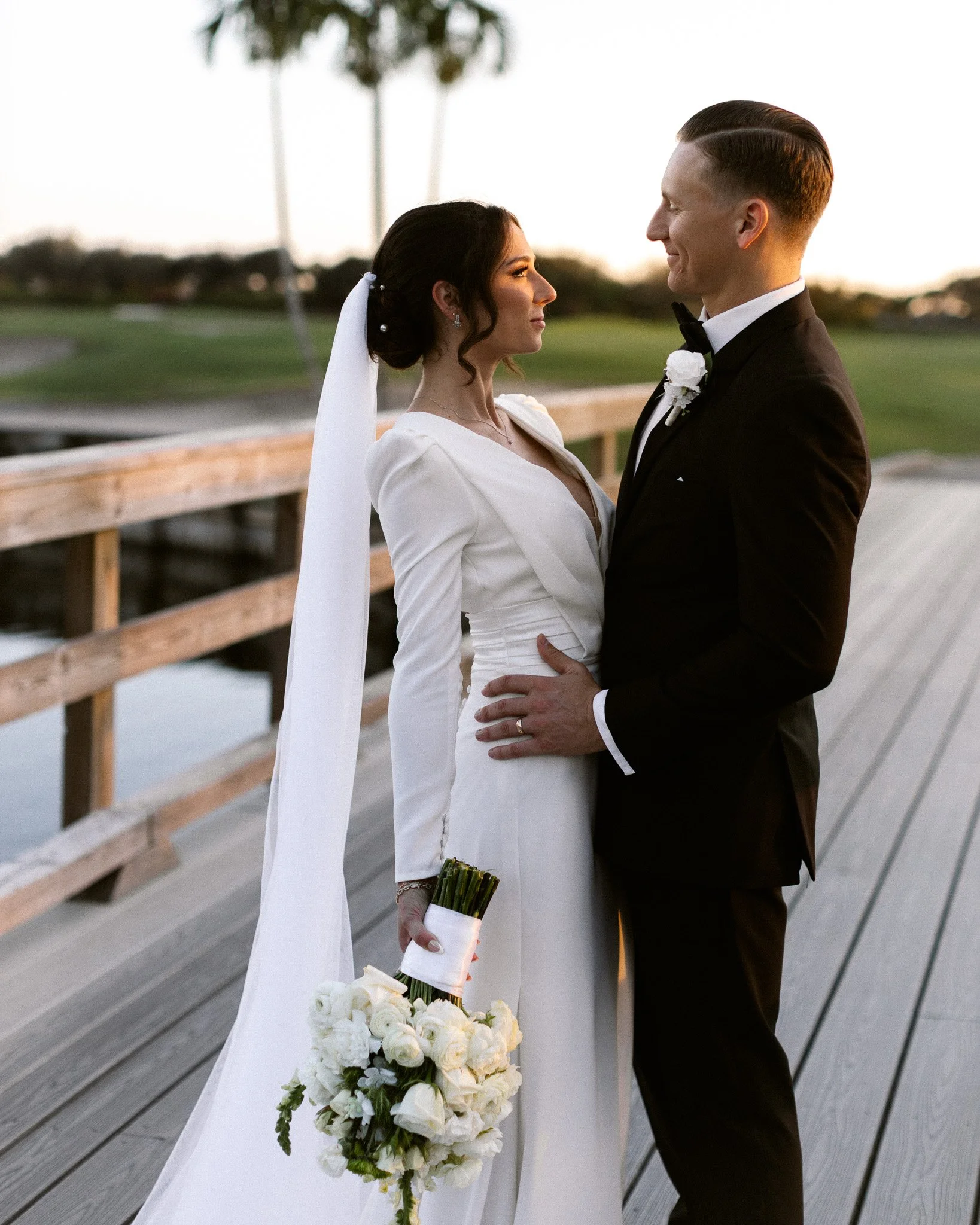 A bride and groom standing close together outdoors on a wooden dock, gazing into each other's eyes during sunset. The bride is dressed in a white gown with long sleeves, holding a bouquet of white flowers. The groom is in a black tuxedo with a white 