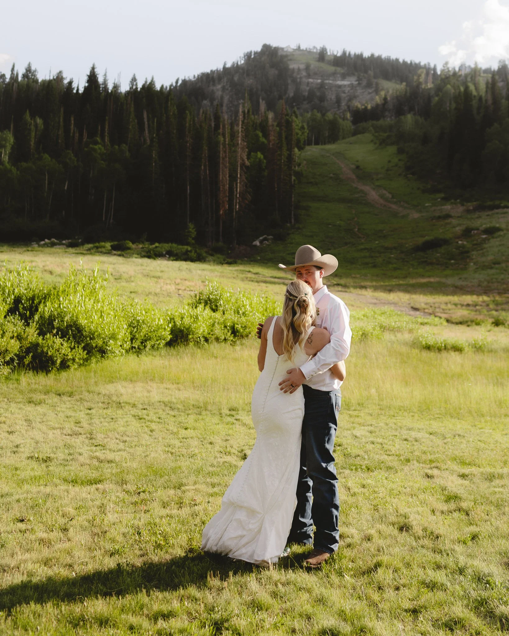 A couple dressed in wedding attire embracing in a lush green field with forested mountains in the background.