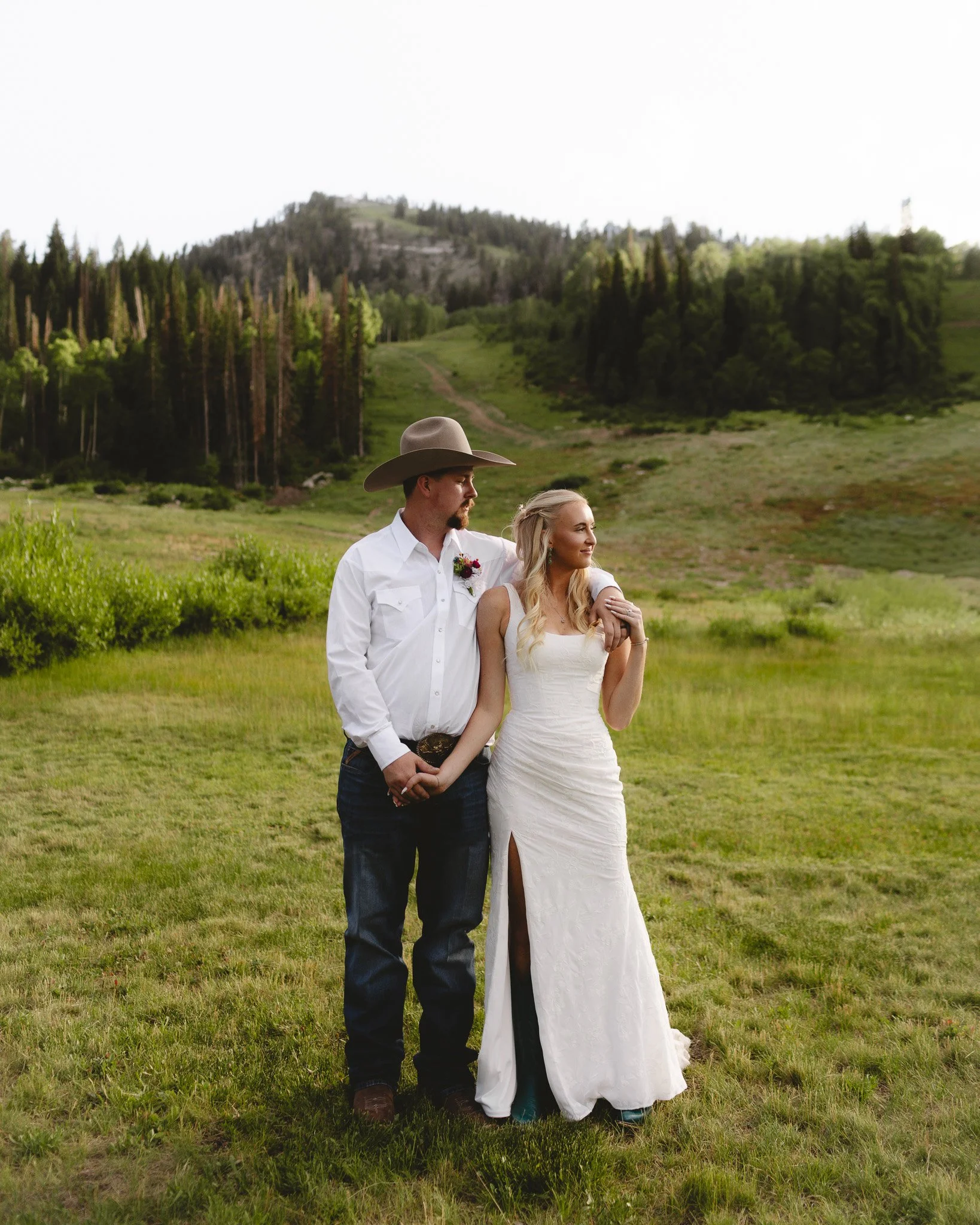A man and woman standing in a green field, with mountains and trees in the background. The man is wearing a cowboy hat, a white shirt, and jeans, while the woman is wearing a white dress. They are standing close, with the man holding the woman's hand and the woman resting her arm on the man's.