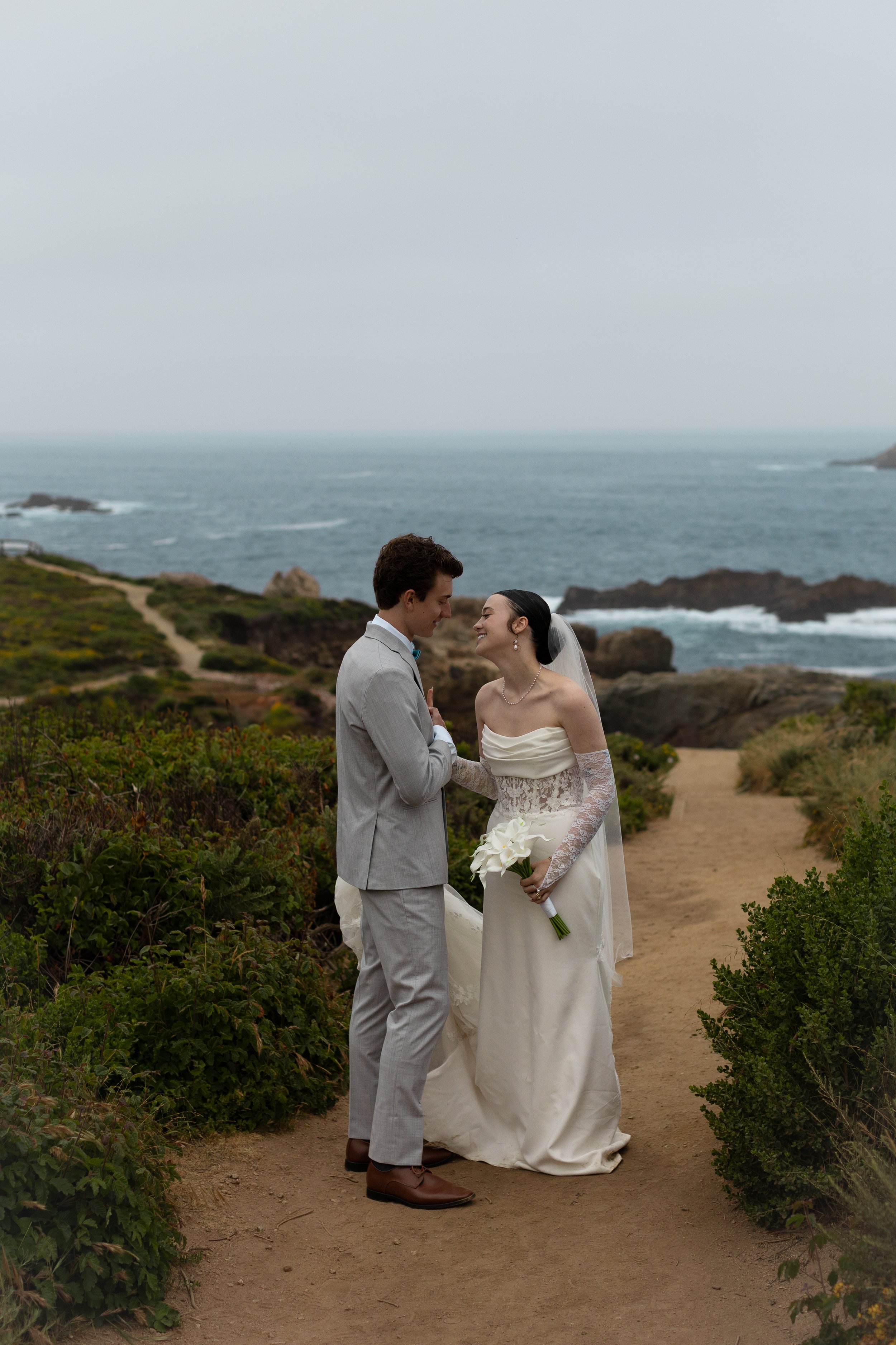 A bride and groom stand on a dirt path near the coast, holding hands and smiling, with the ocean and rocky shoreline in the background.