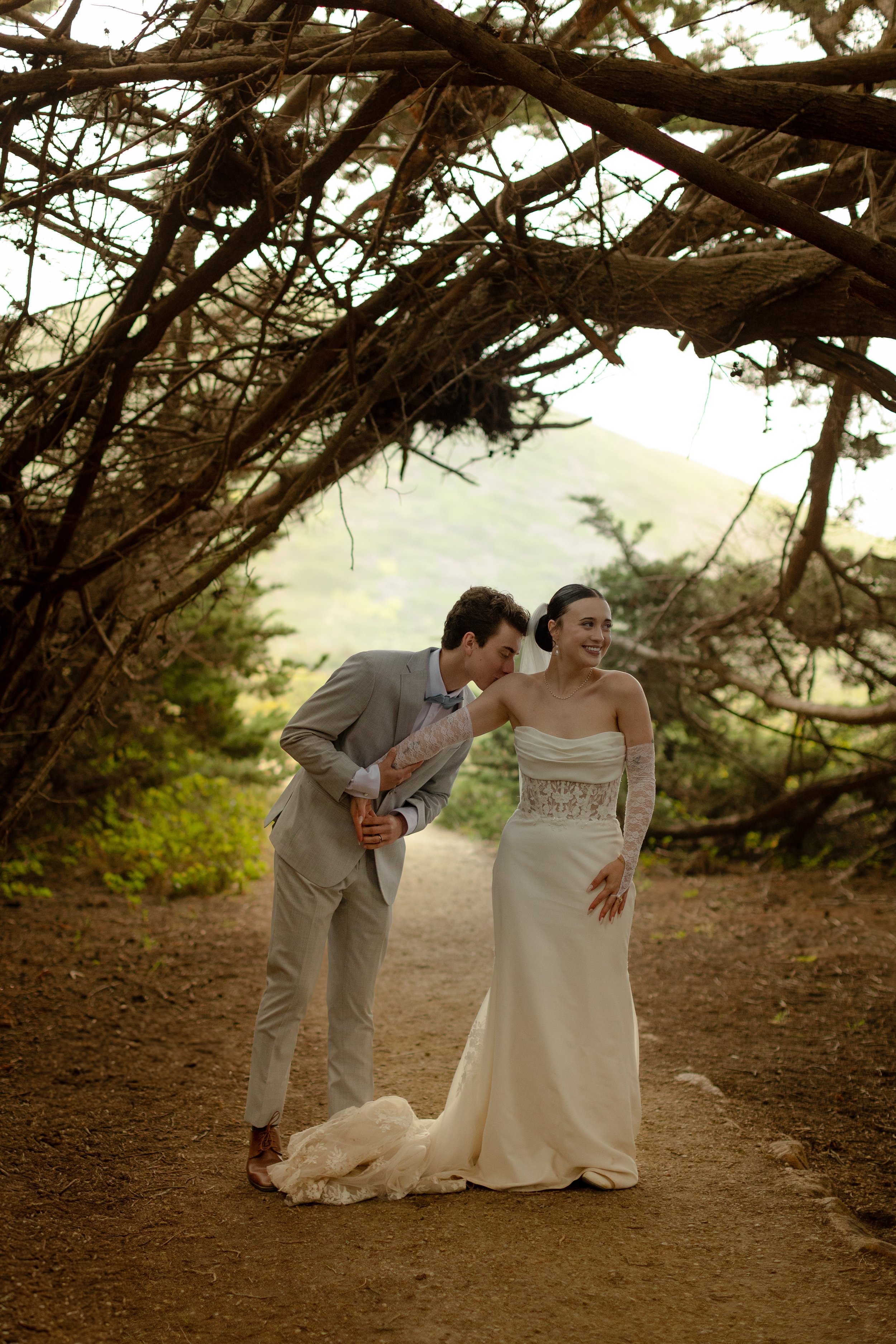 A newlywed couple in wedding attire stands under a natural archway of tree branches and leaves, sharing a joyful moment outdoors amidst greenery, with a scenic mountain background.