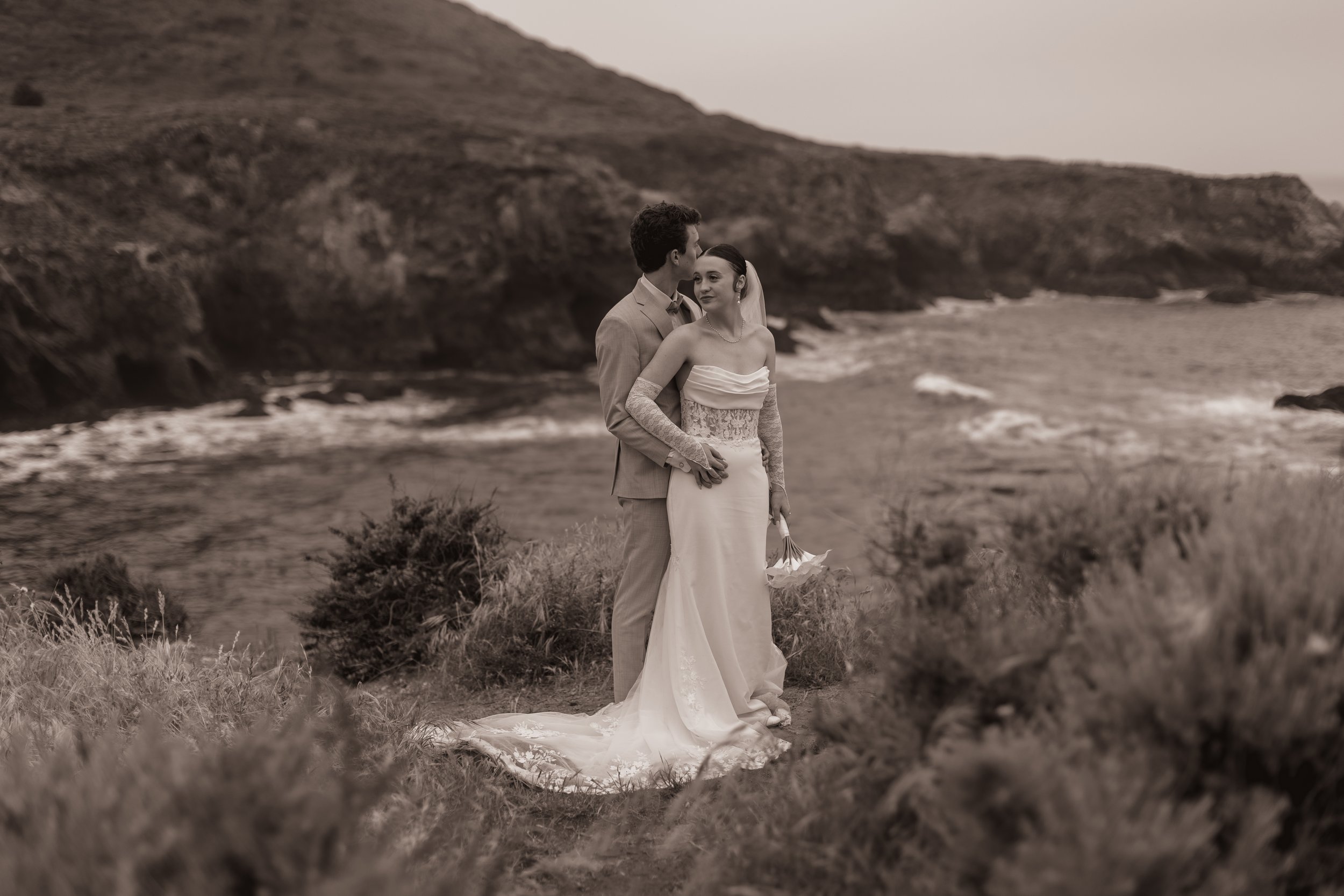 A bride and groom embrace on a grassy hill overlooking the ocean, with rocky cliffs in the background, in black and white.