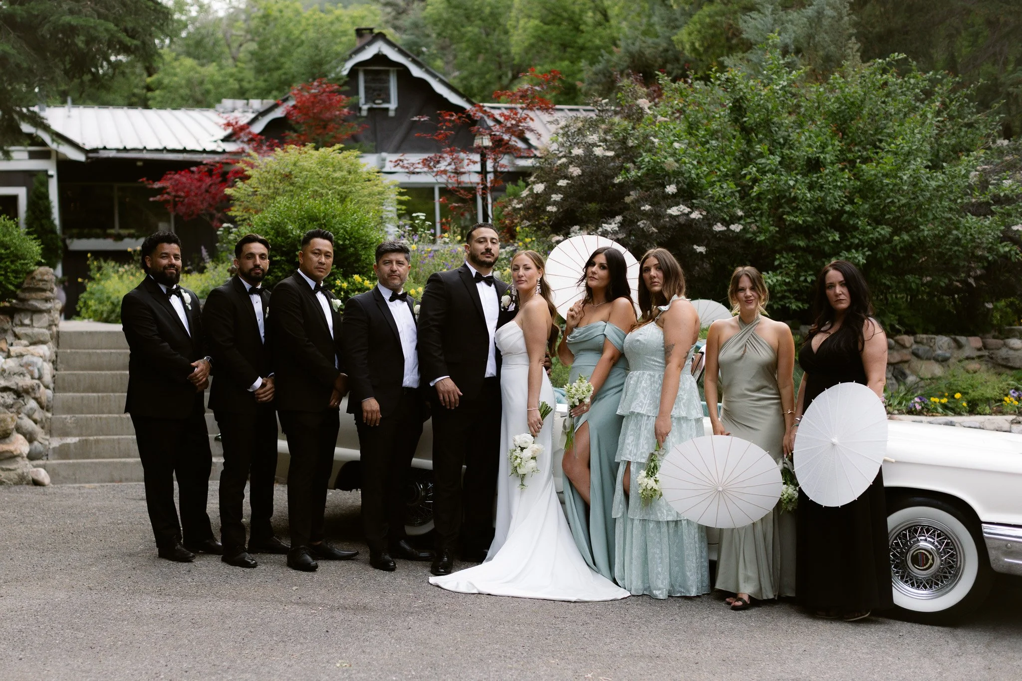 A wedding party with six groomsmen and four bridesmaids stands outdoors in front of a house surrounded by greenery and colorful flowers. The bridesmaids are holding white parasols.