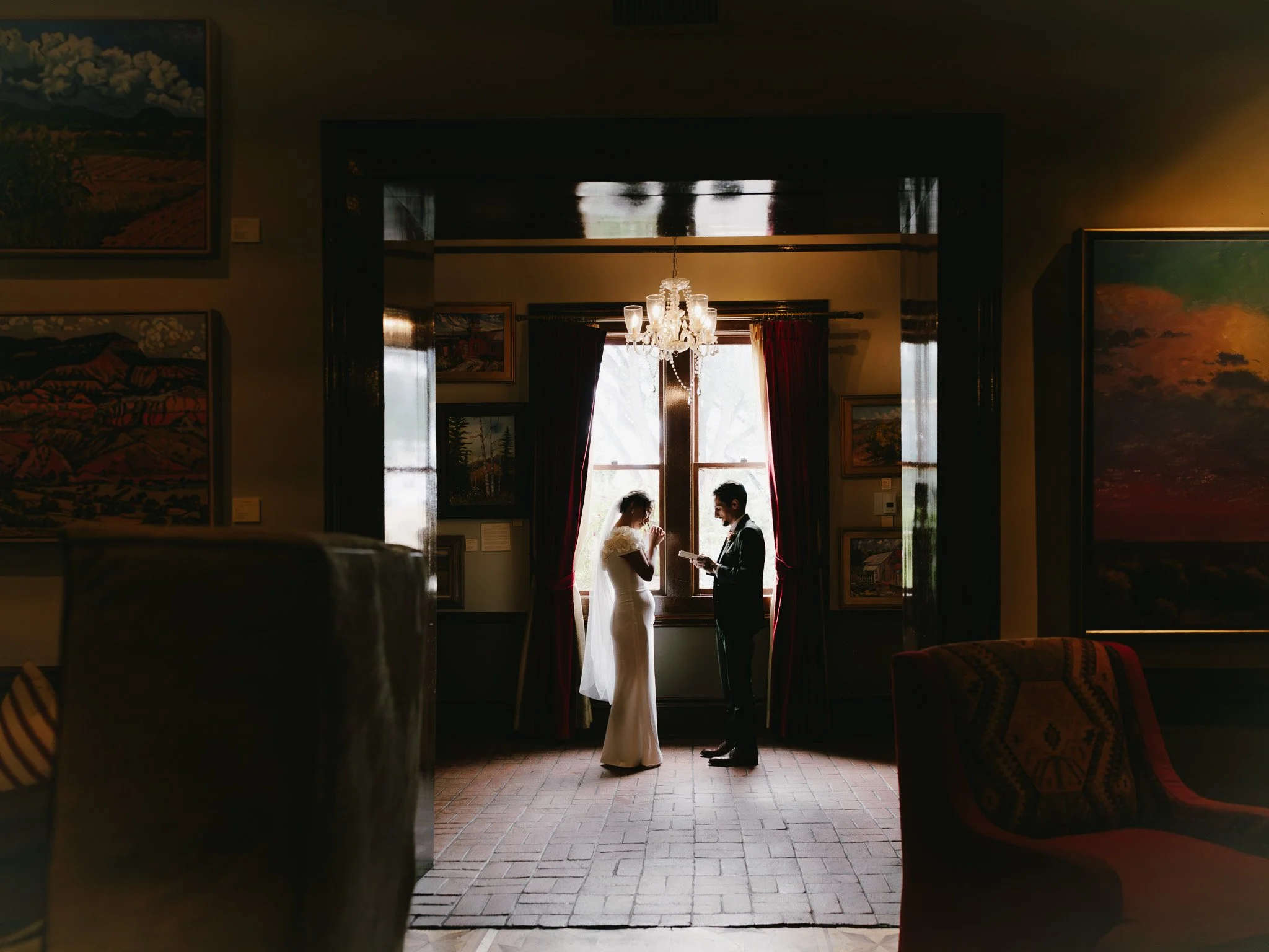 A bride and groom standing in front of a window, exchanging vows during their wedding ceremony inside a room with paintings on the walls and velvet curtains.