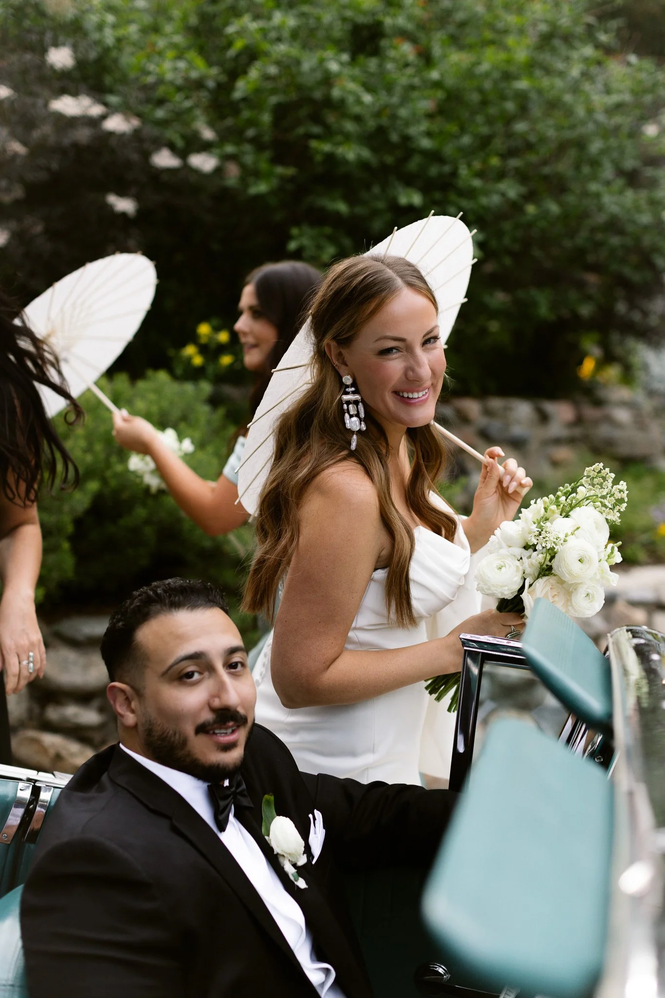 A bride in a white dress with long brown hair, holding a bouquet of white flowers, smiles at the camera. She is sitting in a vintage car with a groom wearing a black tuxedo and bow tie, who is also smiling. Two women in the background hold white para