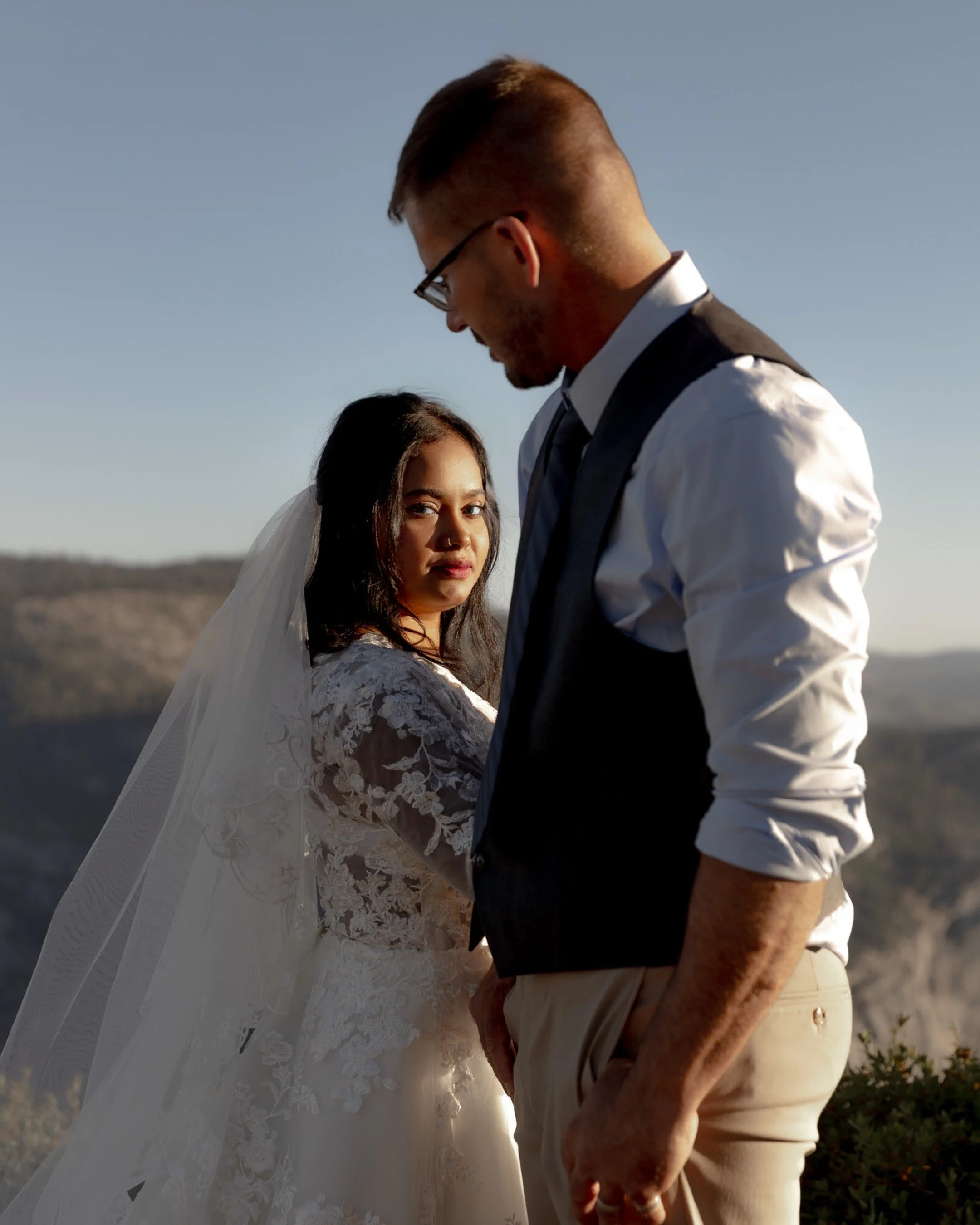 A bride and groom on their wedding day outdoors against a mountain background, with the bride looking at the camera and the groom looking down.