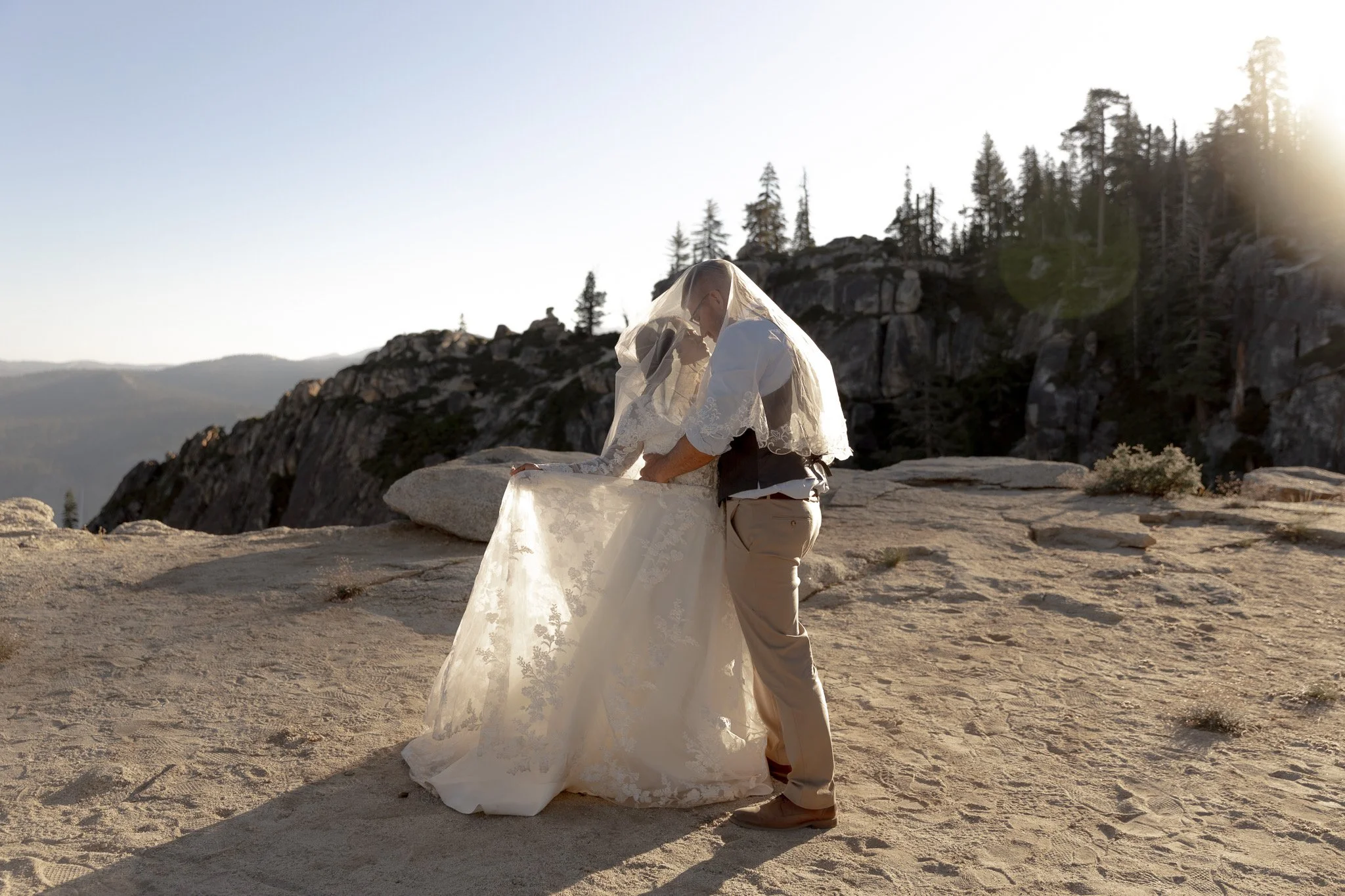 A couple in wedding attire, the bride with a veil and long lace dress, and the groom in khaki pants and a vest, standing on a rocky mountain landscape during sunset.