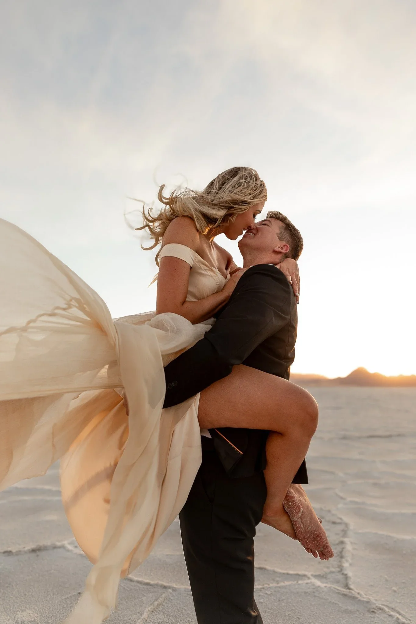 A man lifting a woman in a wedding dress on the beach at sunset, with their faces close together and noses touching at Bonneville Salt Flats, UT. By Jenna Roden.