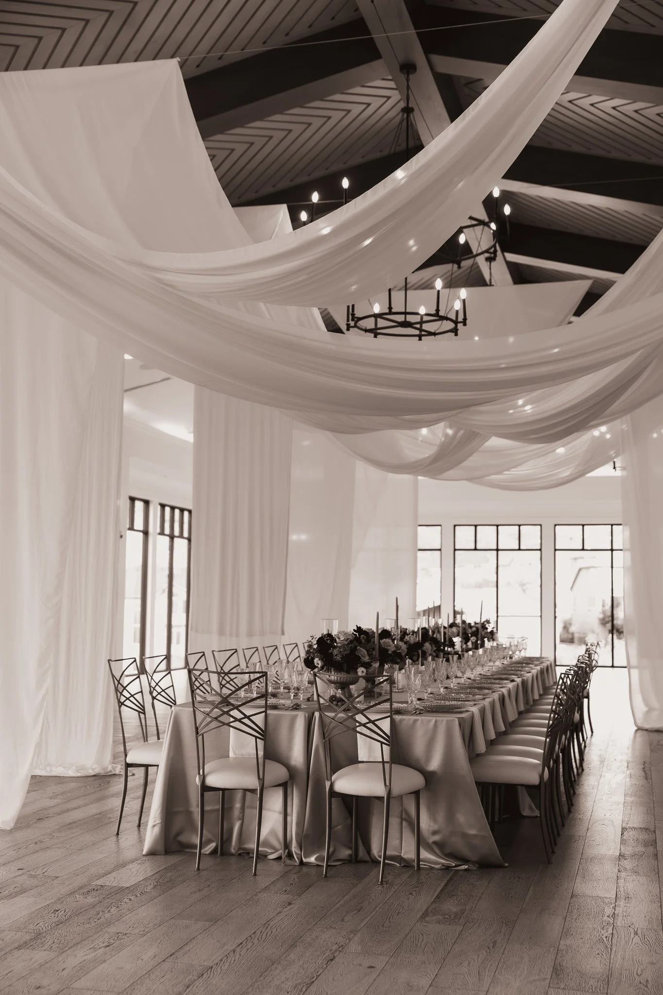 Elegant event space with long dining table set for a formal occasion, decorated with floral centerpieces and candles, white drapery hanging from the ceiling, and large windows in the background.