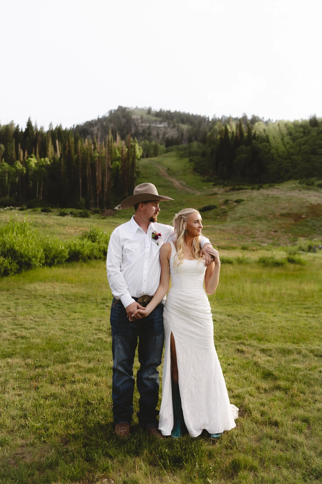 A couple stands outdoors in a grassy field with trees and a mountain in the background. The man wears a white shirt, jeans, and a cowboy hat, while the woman wears a white wedding dress and has long blonde hair. They are holding hands and looking at 