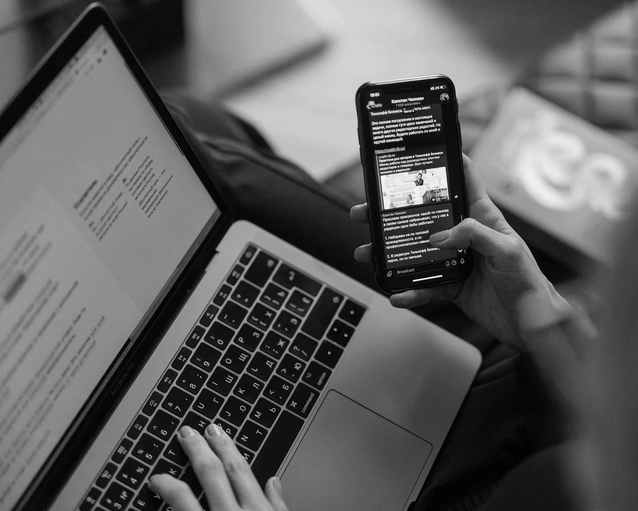 A person using a MacBook Pro laptop with a Russian text document open, while holding a smartphone displaying a chat conversation in Russian. The scene is in black and white.