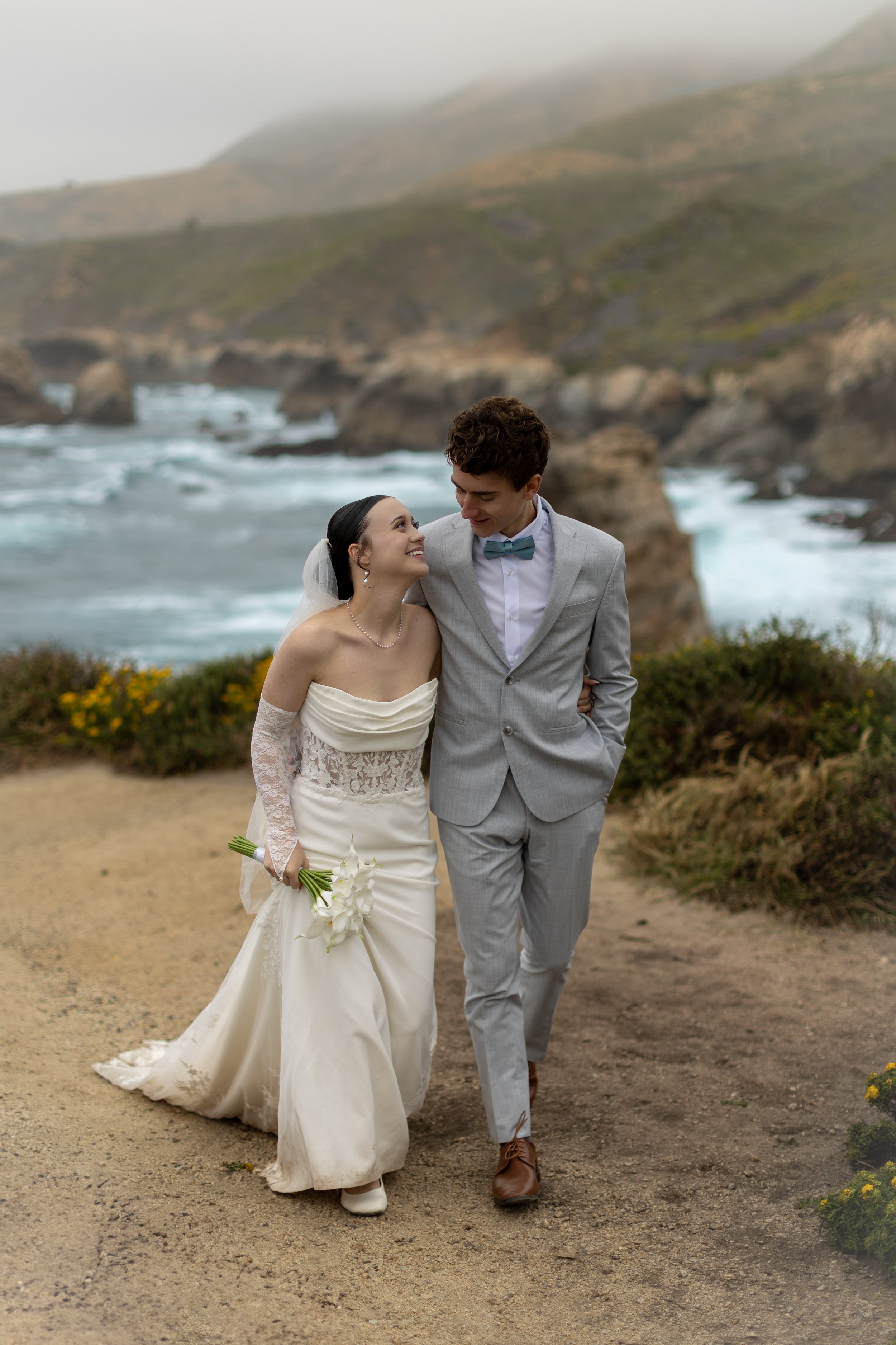 A bride and groom walking together on a coastal trail during their wedding day. The bride is holding a small bouquet of white calla lilies, wearing an off-shoulder wedding dress with lace sleeves, and the groom is dressed in a light gray suit with a