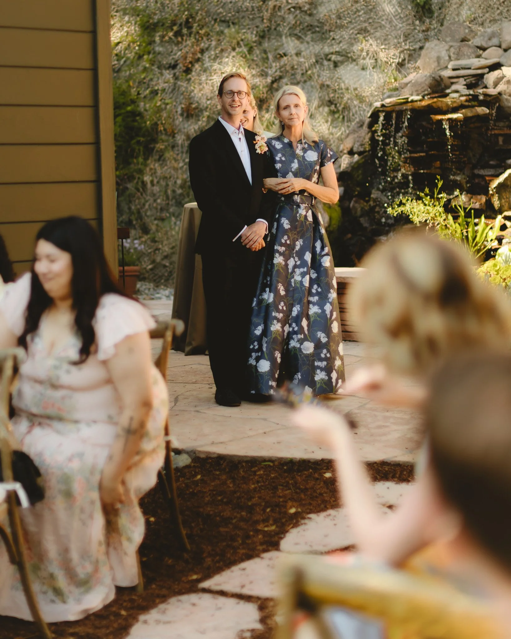 A man and a woman standing outdoors, smiling, during a social gathering. The man is wearing glasses and a suit with a boutonnière, and the woman is in a floral dress. They are near a small waterfall and surrounded by greenery, with other people in th