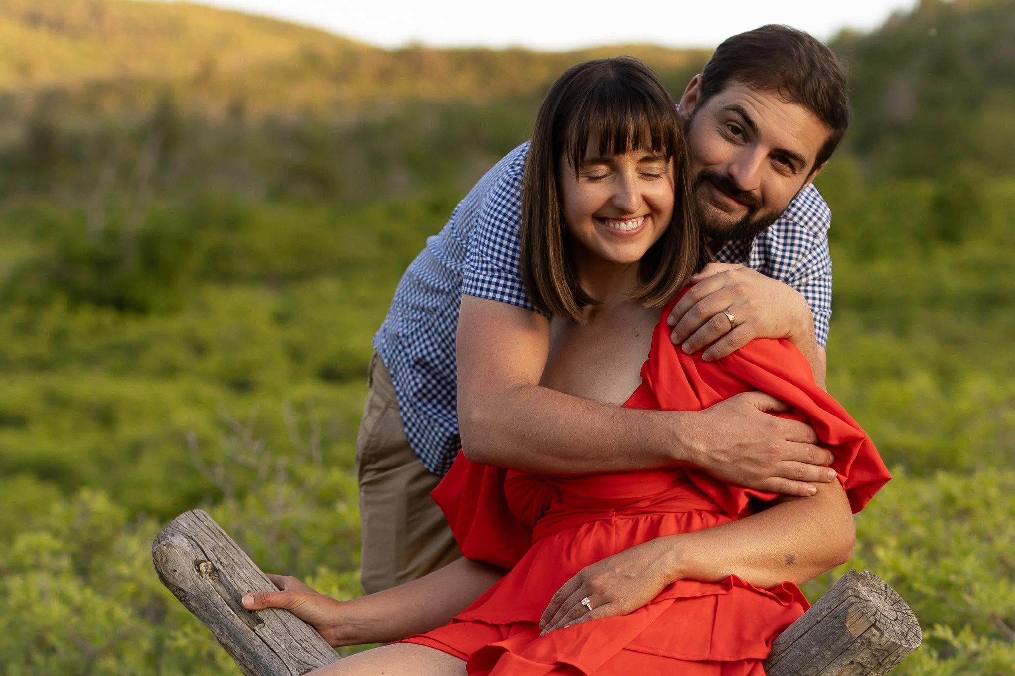 A happy couple is sitting on a log outdoors, embracing each other with trees and a hillside in the background.