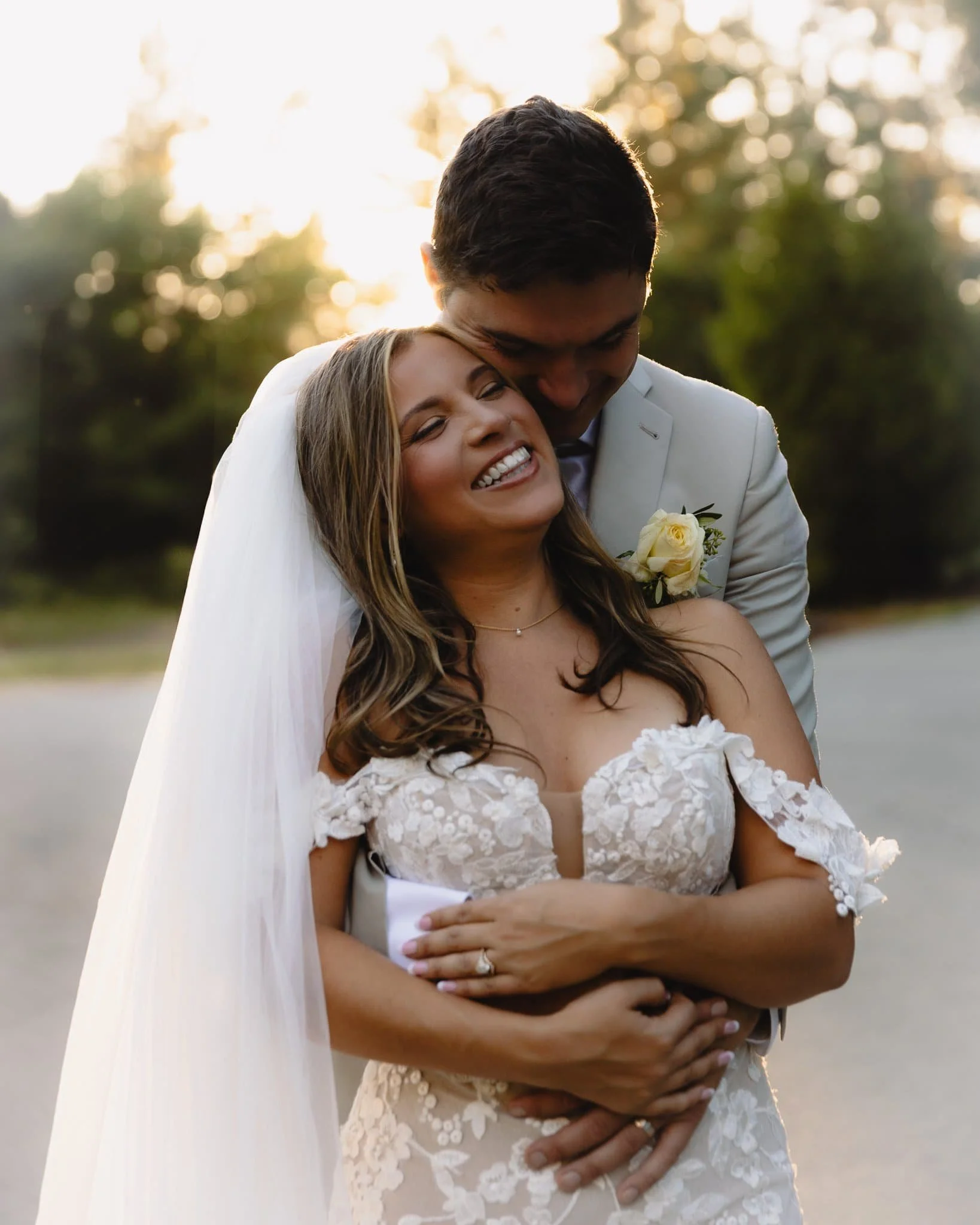 A happy bride and groom embracing outdoors during sunset, with the bride smiling joyfully and the groom kissing her temple.