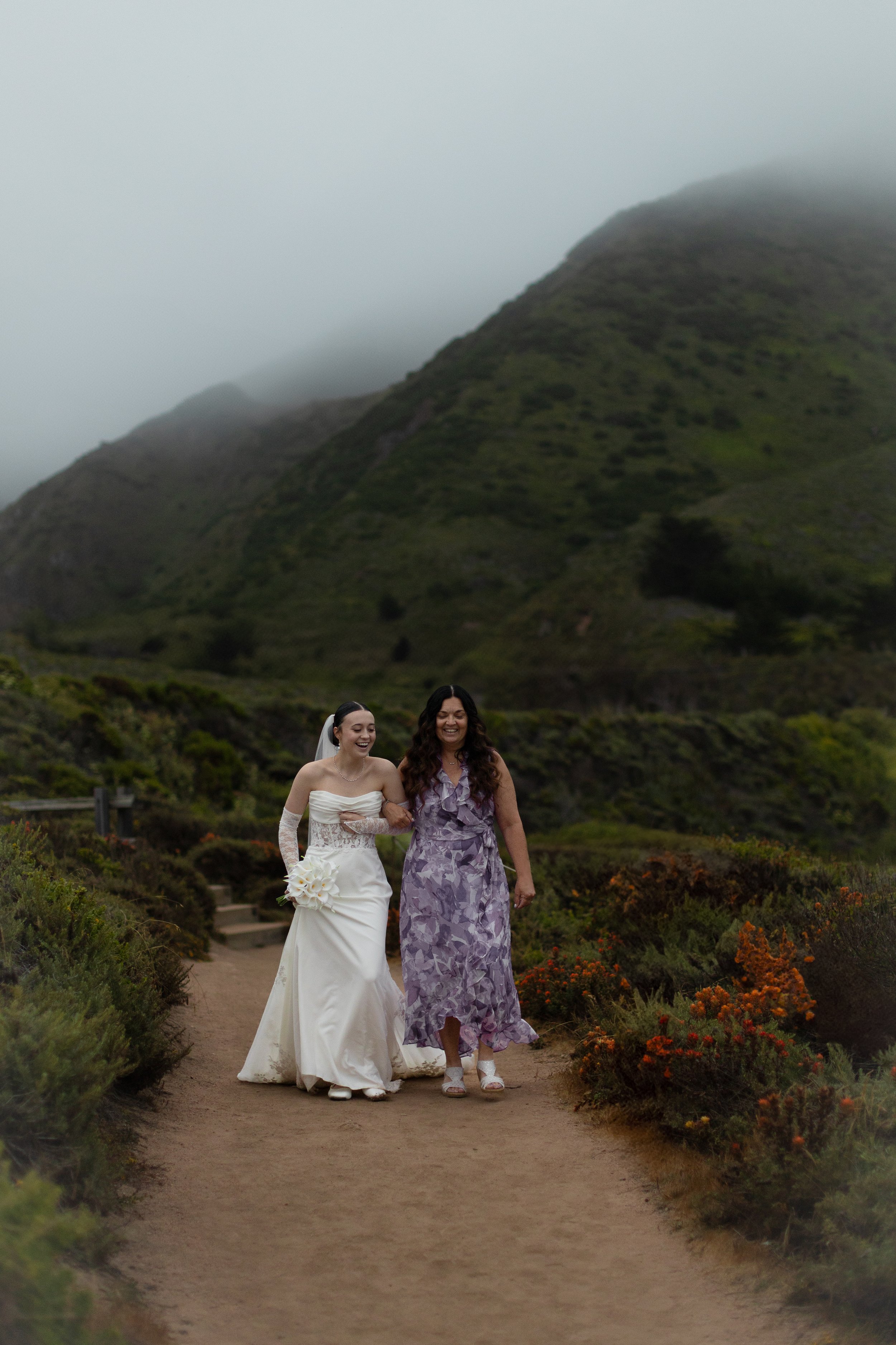 A bride in a white wedding dress holding a bouquet and with a veil, walking arm-in-arm with a woman in a purple dress on a dirt path surrounded by greenery and flowers, with foggy mountains in the background.