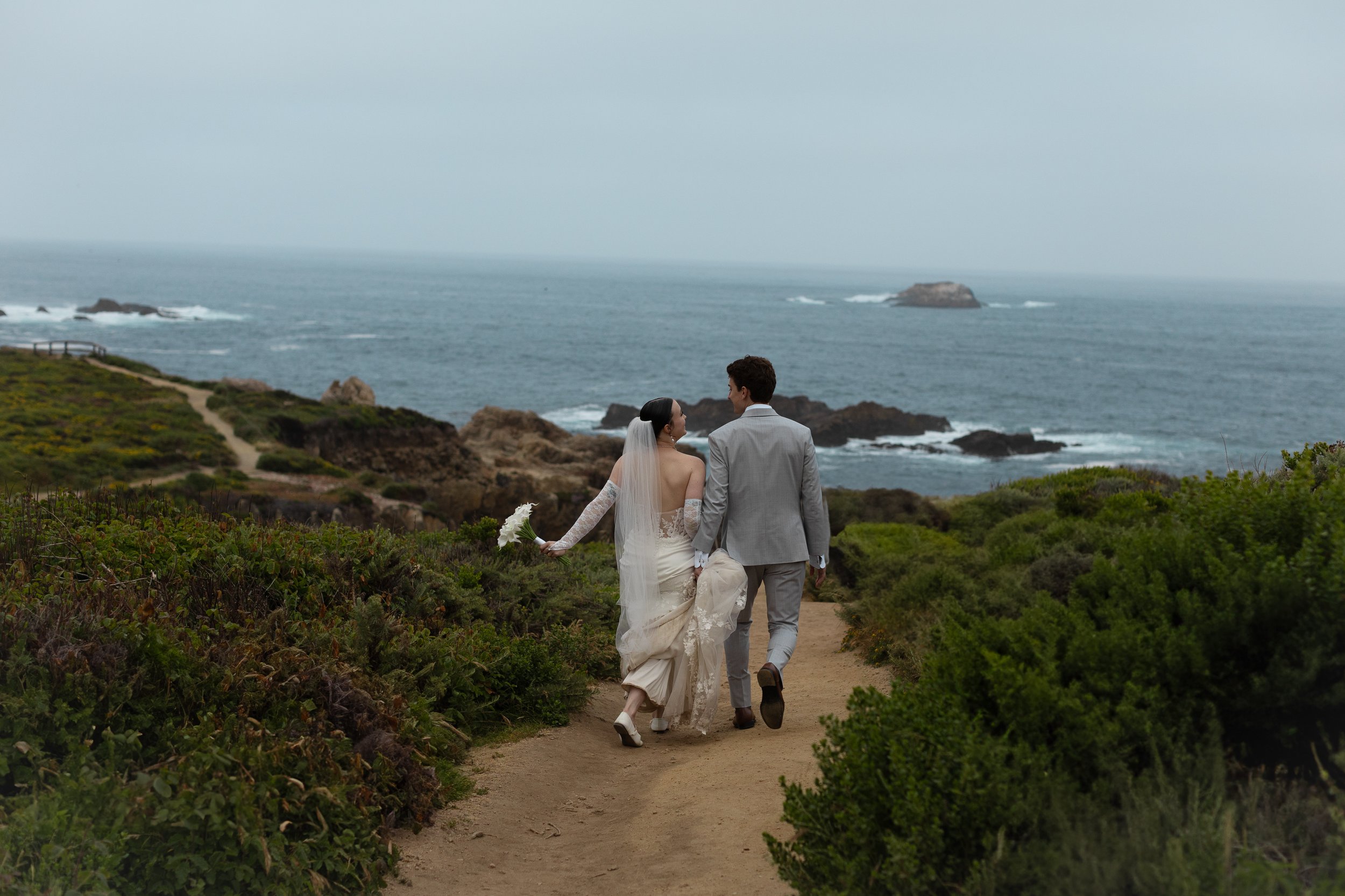 A bride and groom walking along a dirt path near the coast, with ocean and rocky shoreline in the background, holding hands and facing away from the camera.