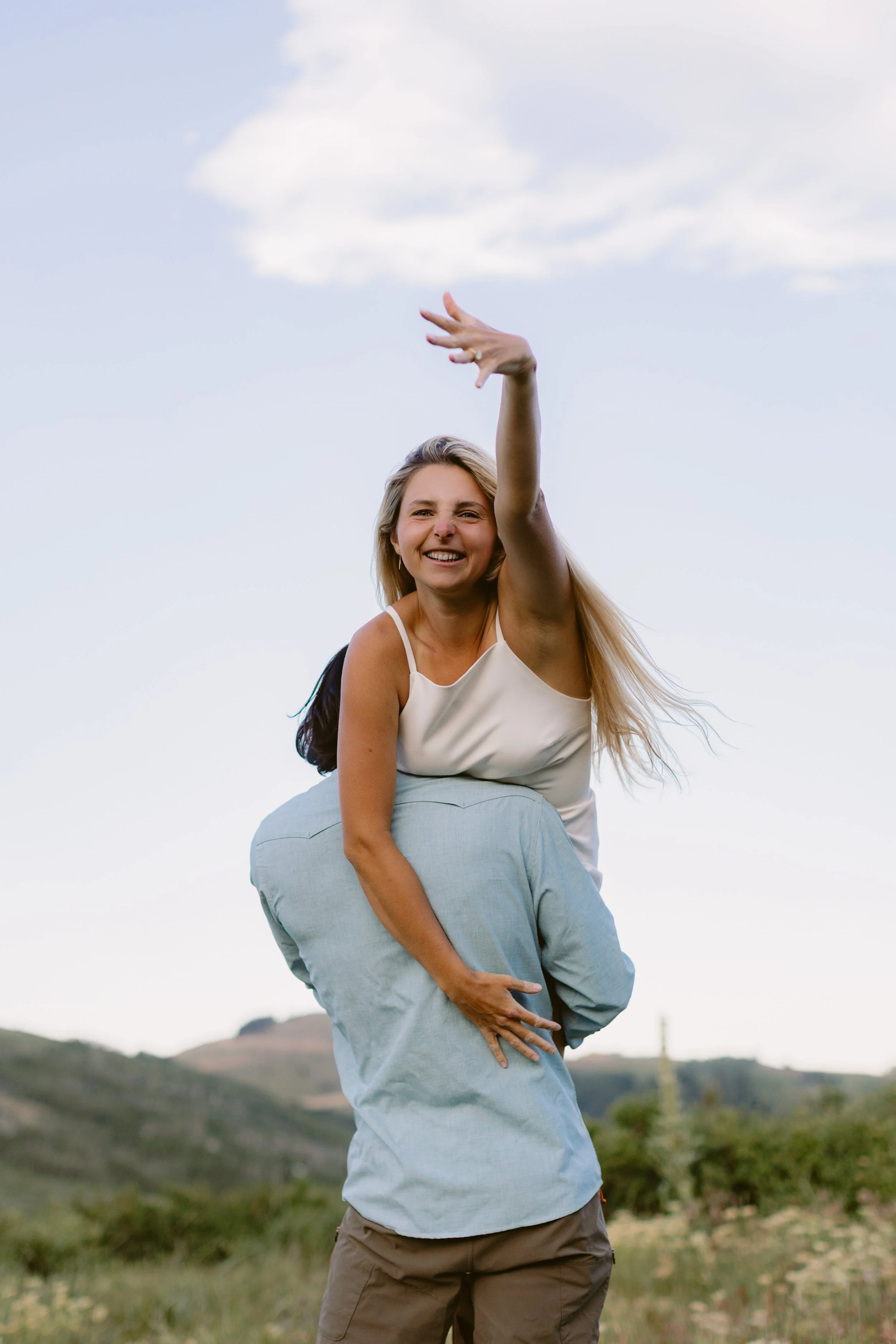 A woman smiling and leaning over a man's shoulder, with her arm raised in a playful gesture, outdoors in a grassy area with hills and a blue sky with clouds.