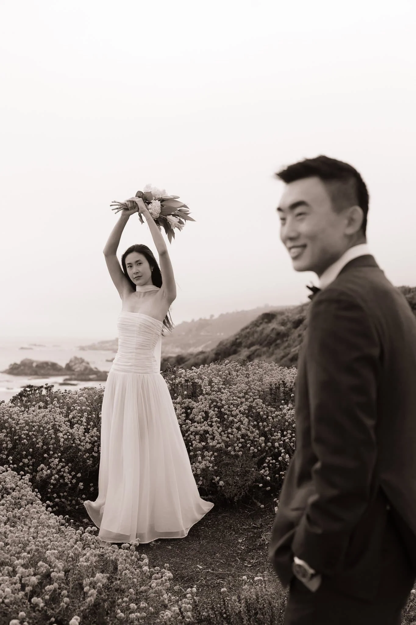 editorial wedding photographer capturing bride with bouquet raised at Garrapata State Park coastal cliffs with groom in foreground