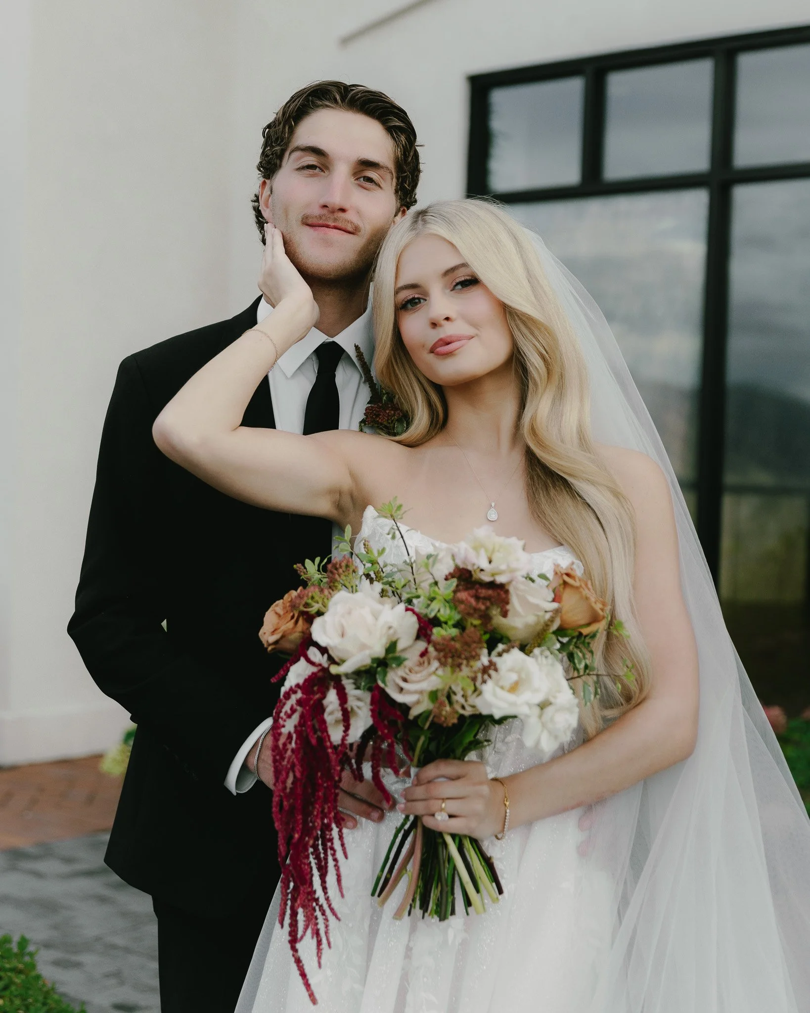 Bride and groom standing together outside, with the bride holding a bouquet of flowers, the groom wearing a black suit and tie, and the bride in a white wedding dress with a veil.