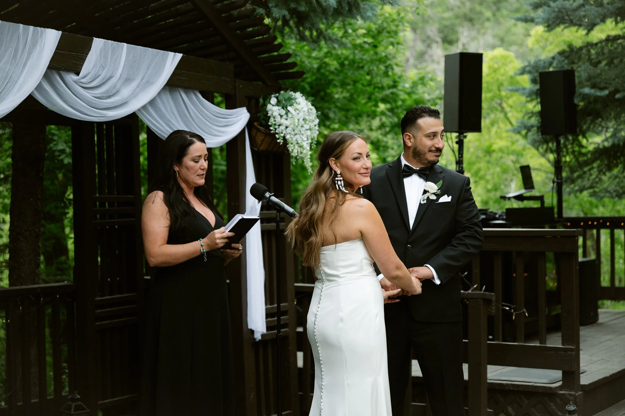 A bride and groom standing in front of an officiant during an outdoor wedding ceremony with greenery in the background.