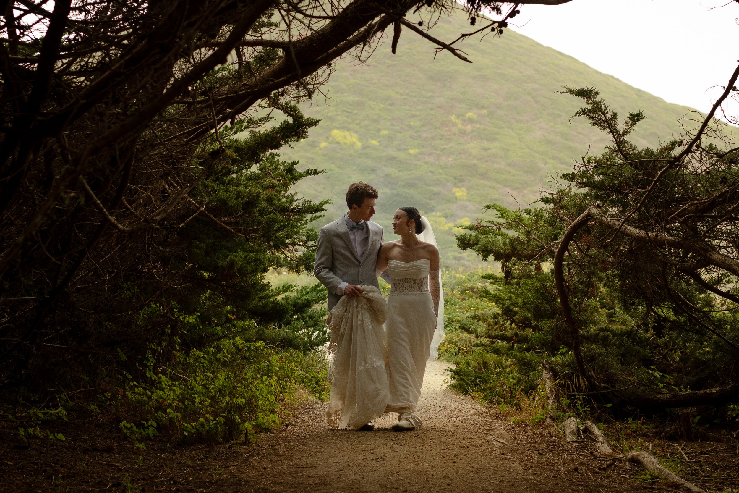 A bride and groom walking together on a dirt path surrounded by trees, with green hills in the background, during daytime.