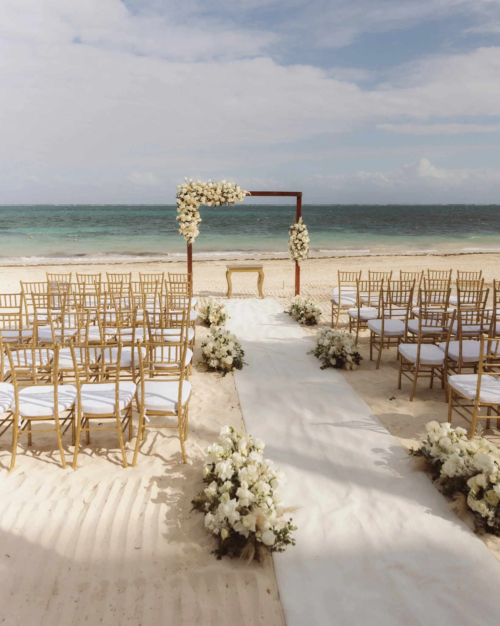 Beach wedding ceremony setup with gold chairs, floral arrangements, and a wooden arch with flowers, on a sandy beach facing the ocean.