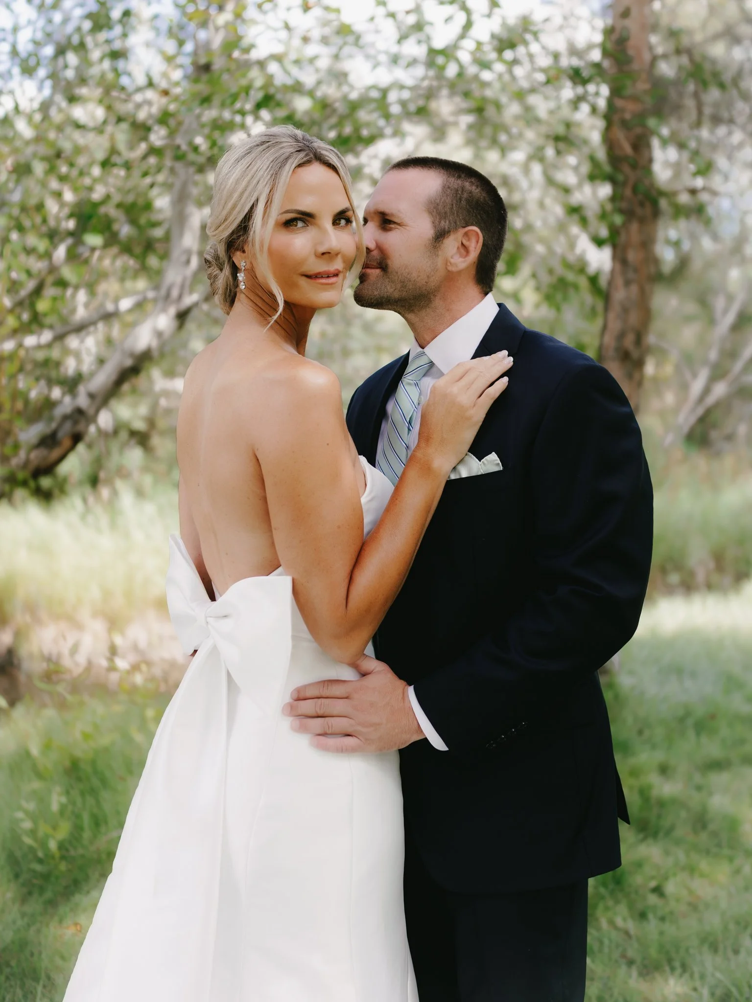 A bride and groom standing outdoors, embracing each other, with trees and greenery in the background.