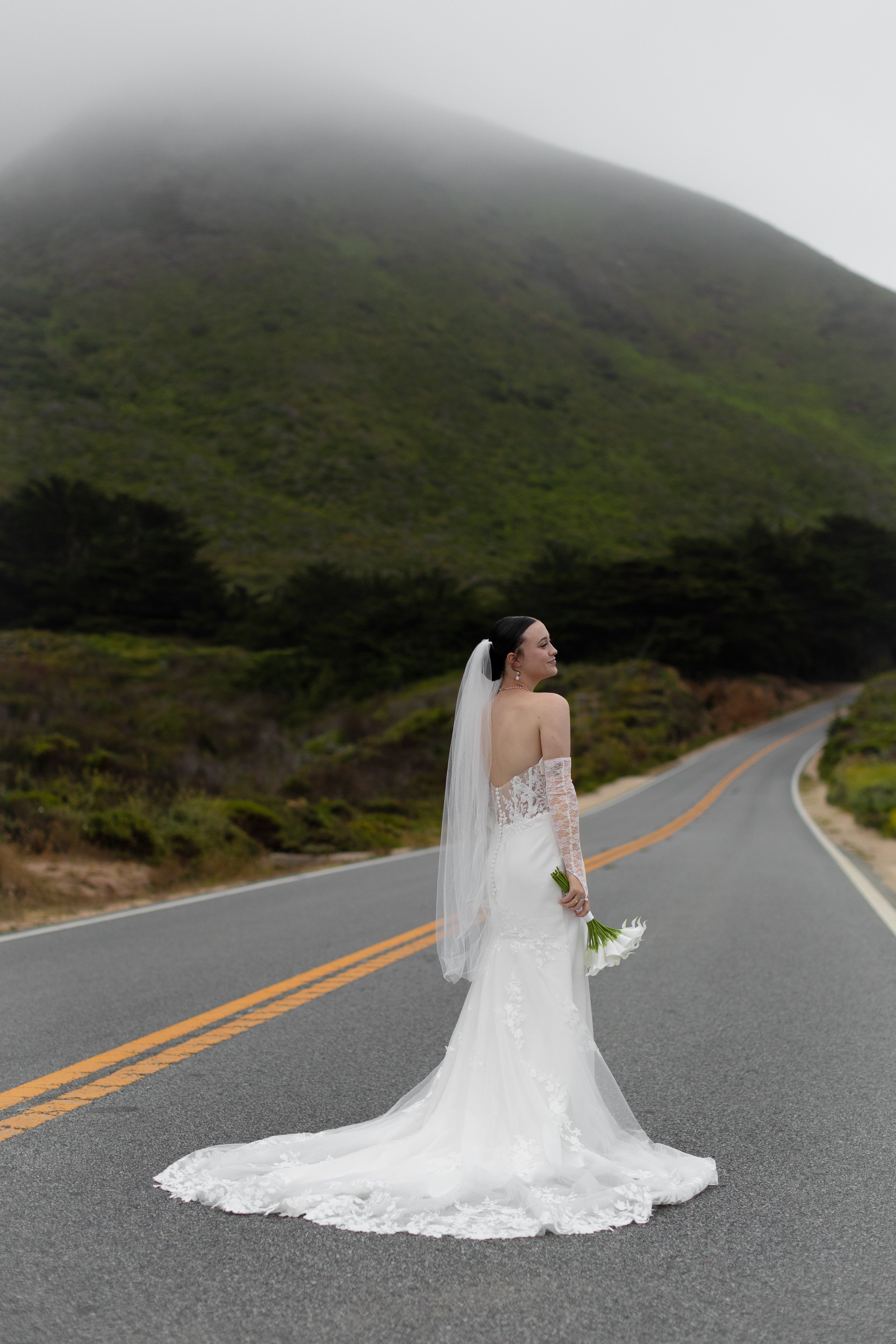 Bride in a white wedding gown and veil holding a bouquet standing on an empty winding mountain road with green hills and fog in the background.